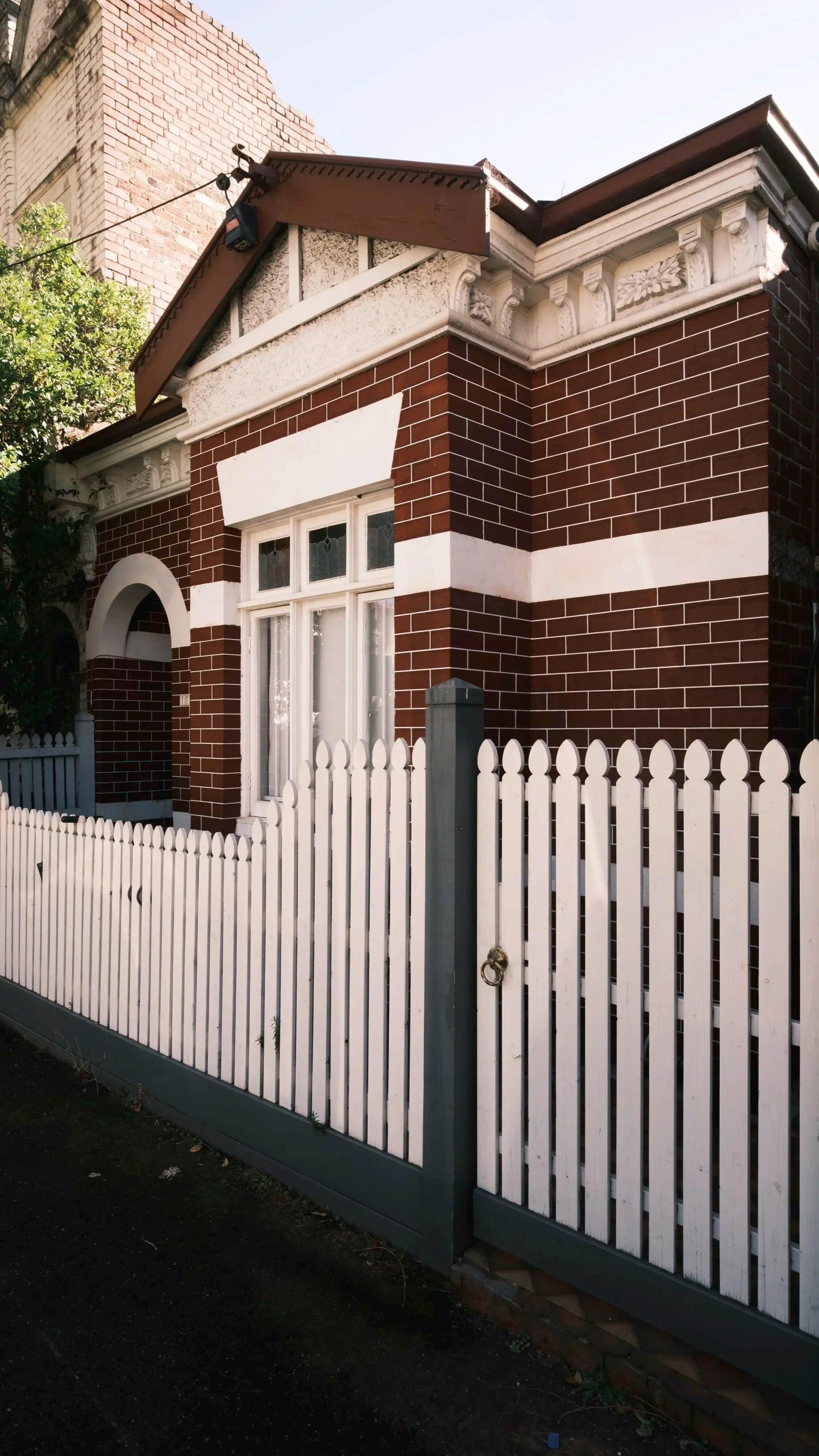 A brick house with a white picket fence in front and a large window, with decorative convex windowpanes, on the right side of the house. There is a section of the house's roof visible at the top of the image.