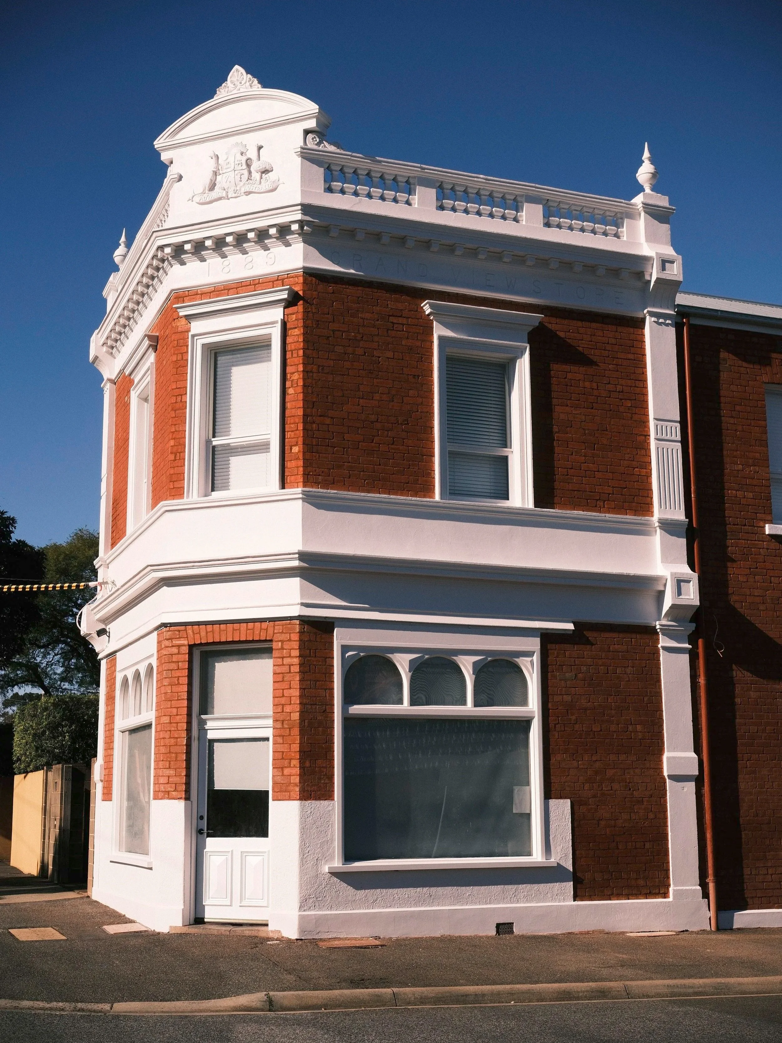 A corner building with a red brick facade and decorative white trim around the windows and roof. The building has large ground-floor windows and a white door. The top portion has ornate white architectural details, including a crest or emblem and balustrades, against a clear blue sky.