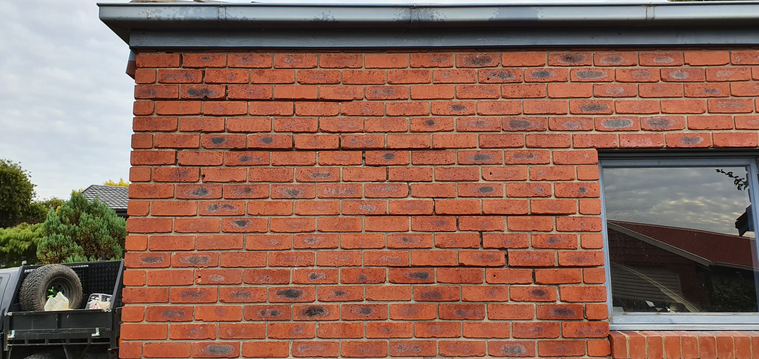 Close-up of a red brick exterior wall of a house with a large window and a gray roof overhang. Part of a yard with trees and a trailer is visible in the background.