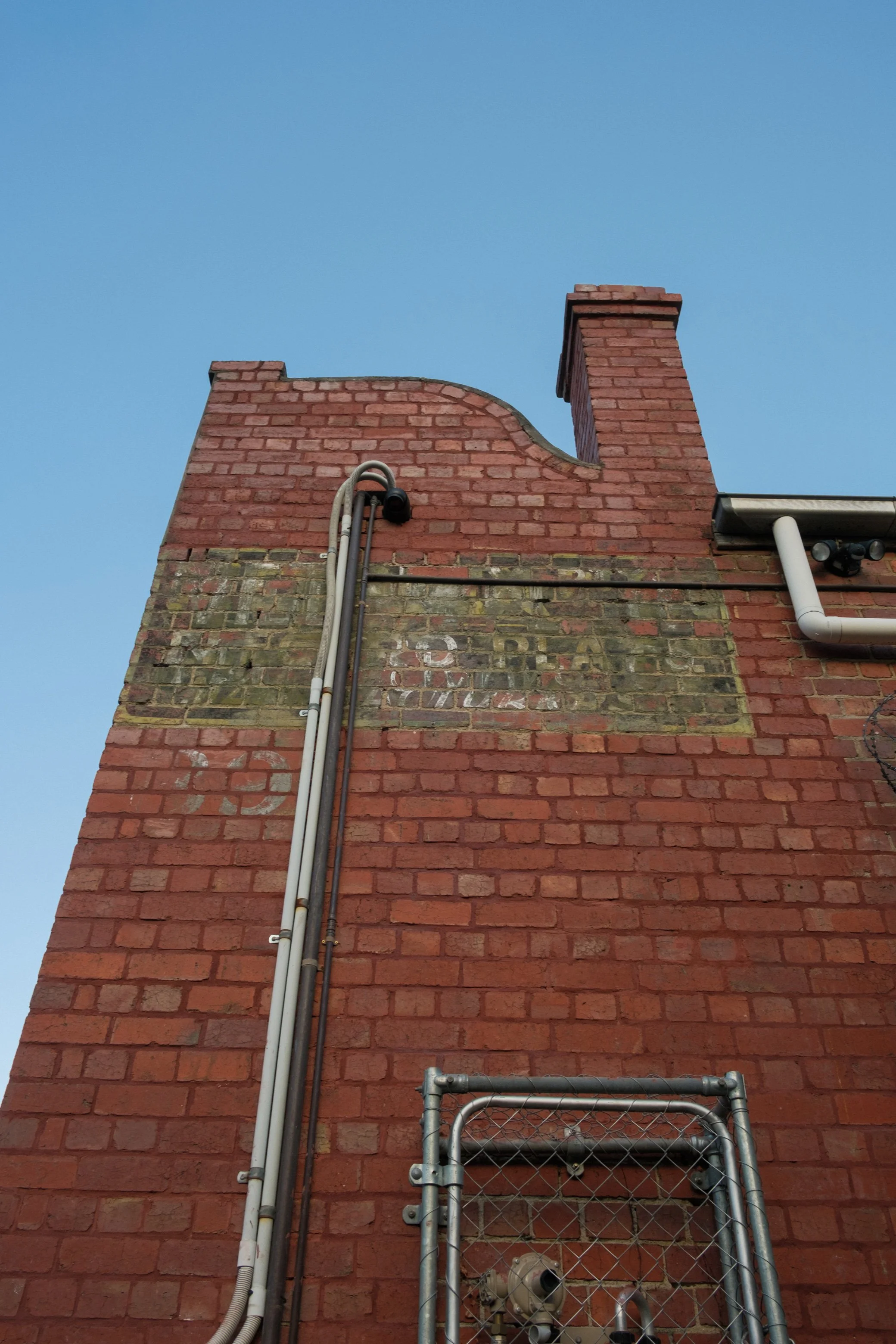 Upward view of a brick building wall with a chimney and utility pipes against a clear blue sky.