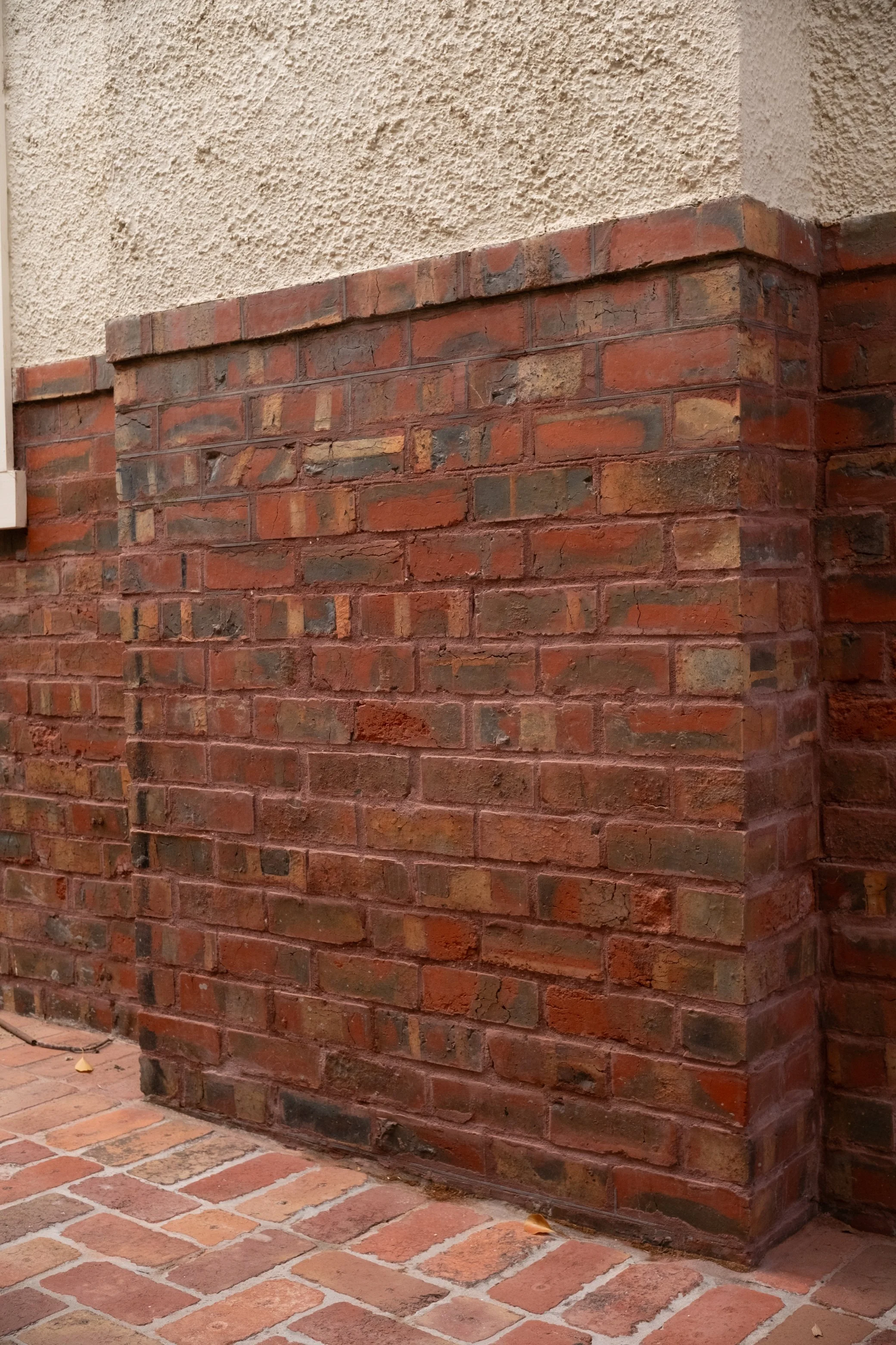 Close-up of a brick corner wall with a textured beige stucco wall above, and brick pavement at the base.