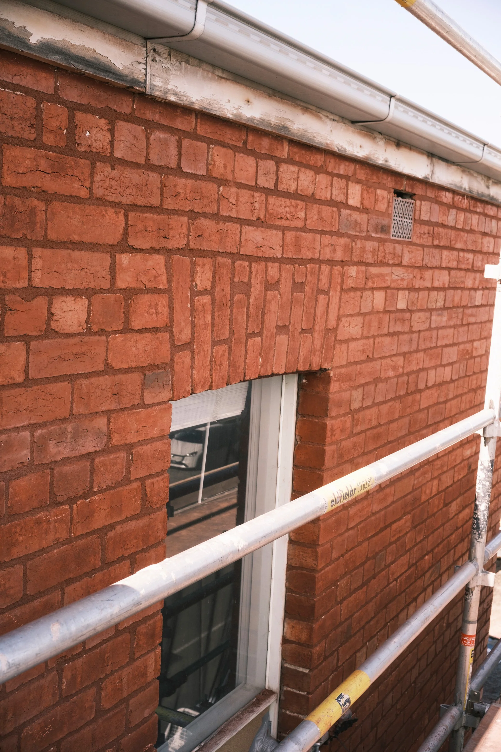 A window in a brick wall with scaffolding in front. The brick wall shows signs of weathering and peeling paint near the top.