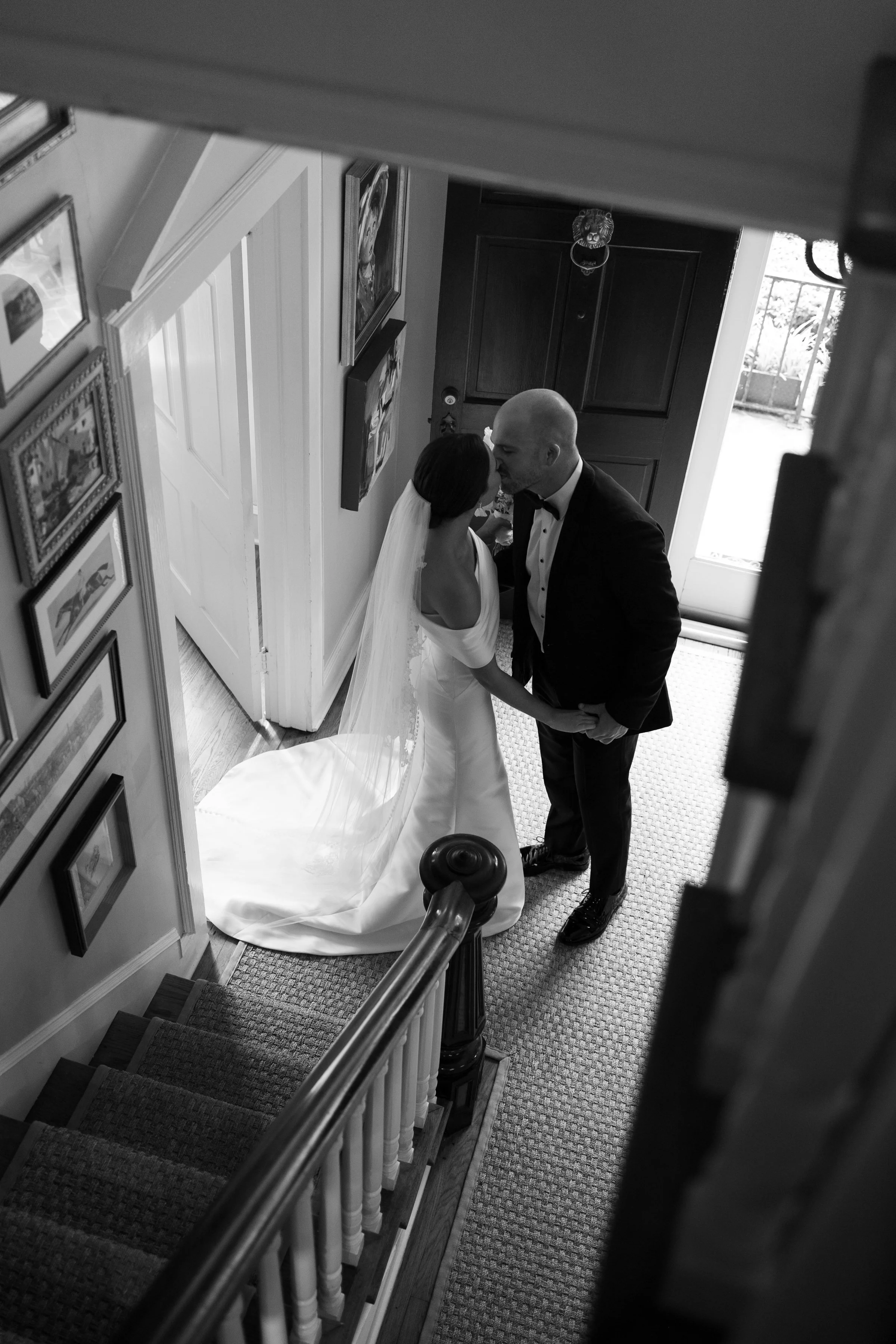 A black and white photo of a bride and groom standing at the top of a staircase, holding hands and about to kiss, in a home with framed pictures on the wall and a window letting in natural light.