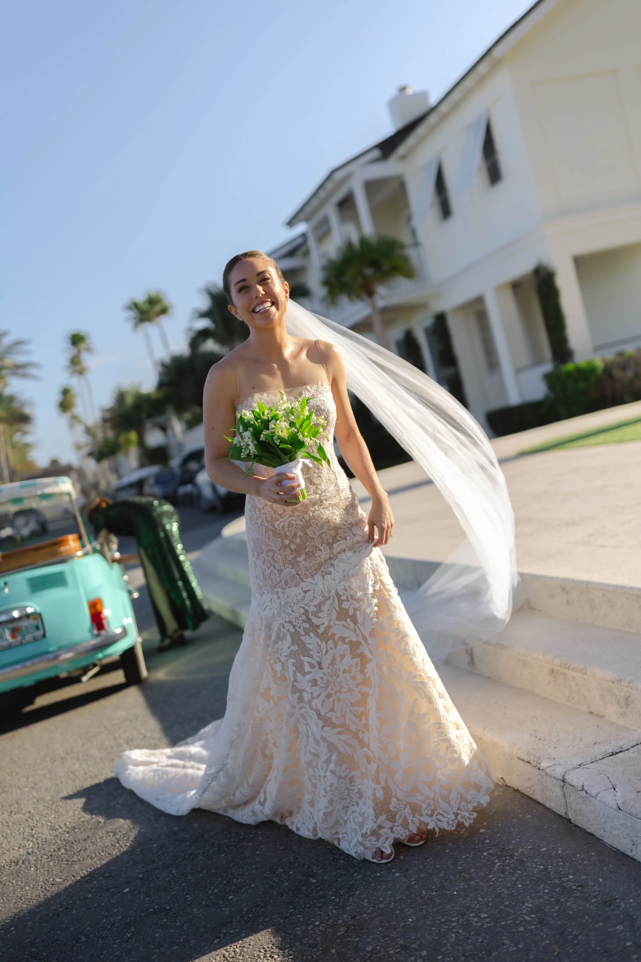 A joyful bride in a white lace wedding dress holding a bouquet of green flowers, smiling in front of a white house and palm trees, with a vintage car nearby on a sunny day.