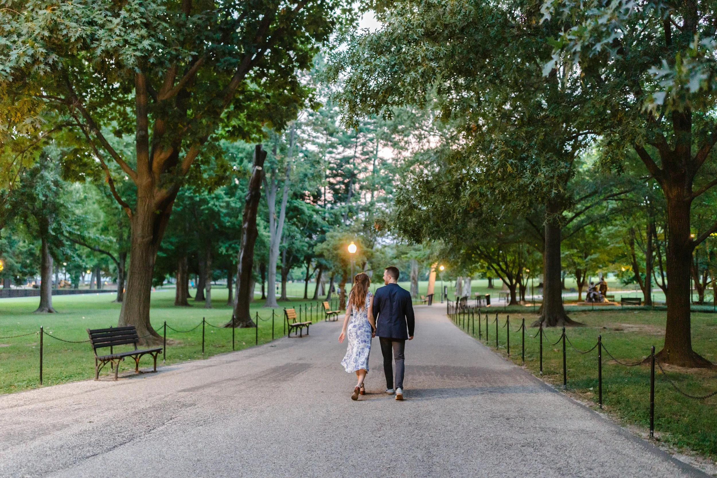 A couple walking hand in hand along a park path at dusk, surrounded by trees and park benches.