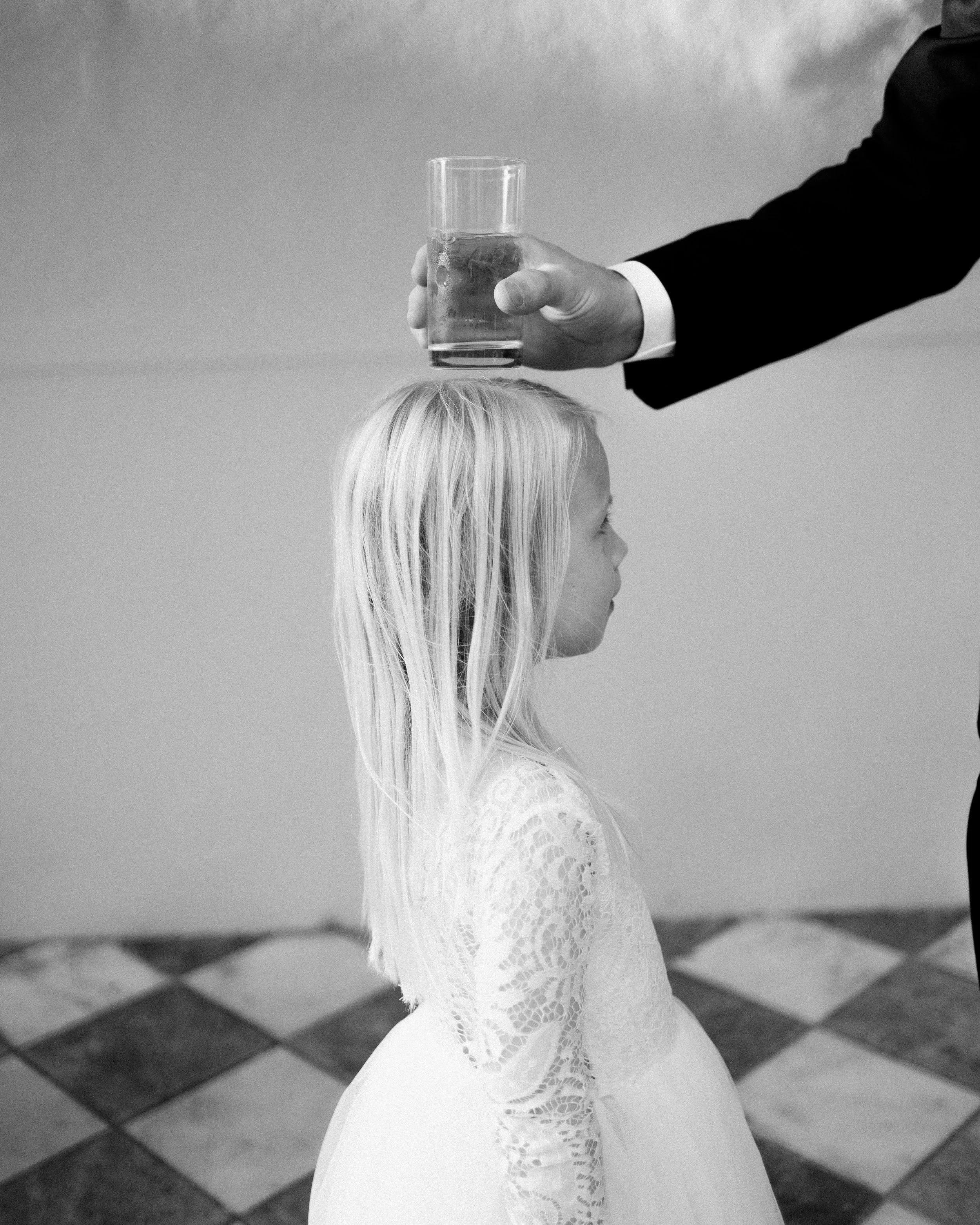 A young girl with long blonde hair and a white dress standing sideways as an adult extends a hand holding a glass of water over her head. The adult is wearing a suit jacket.