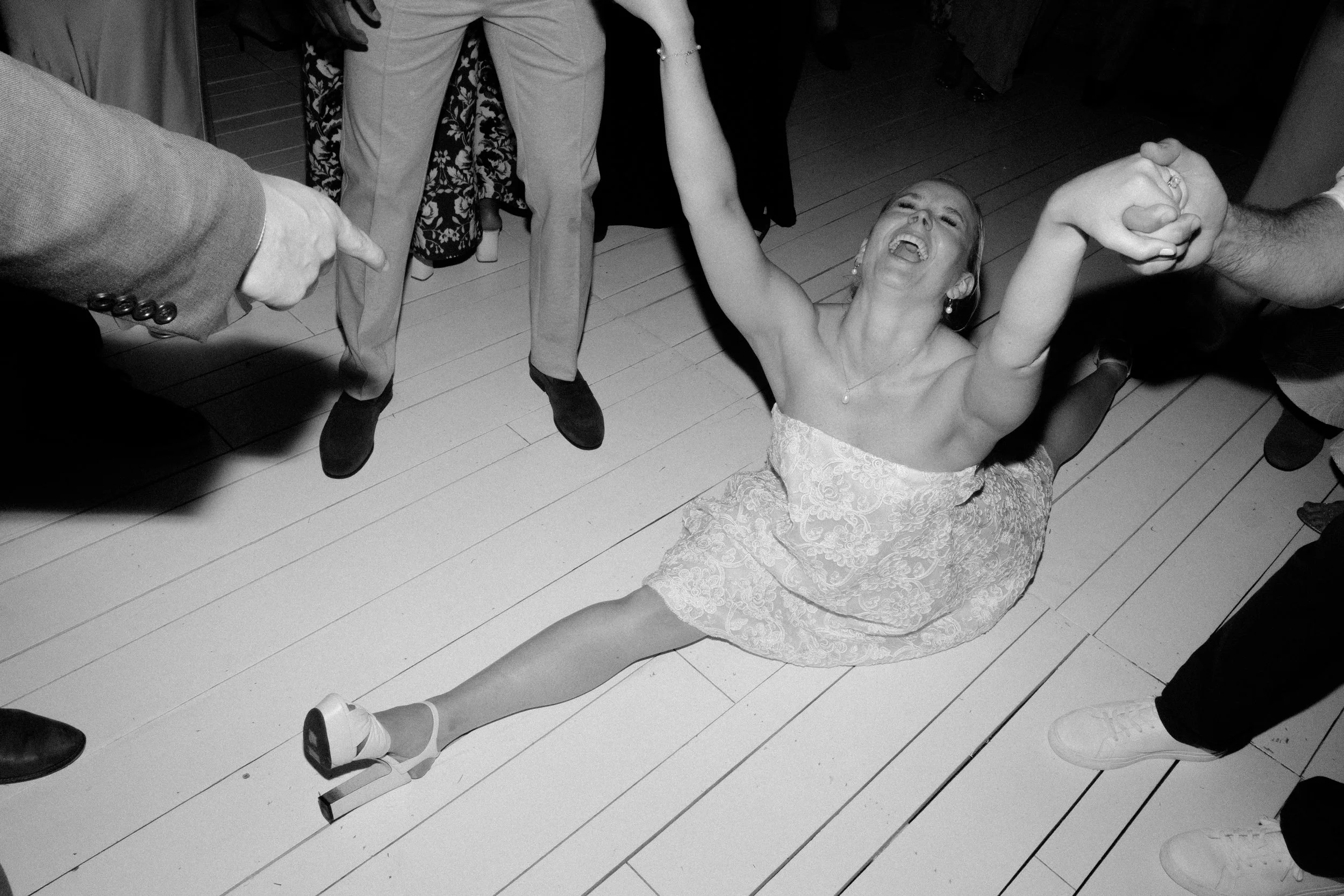 Woman in a strapless dress sitting on the floor, smiling and laughing, holding hands with two people, surrounded by others at a social gathering or celebration.