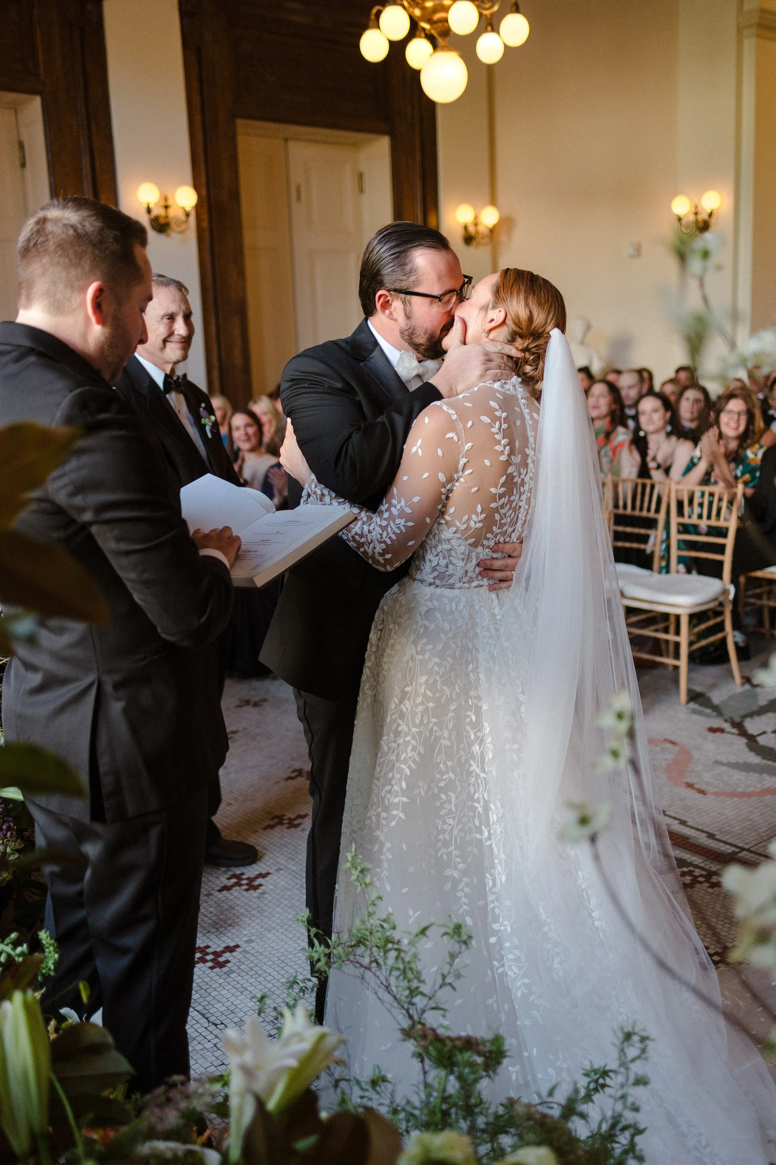 A bride and groom share a kiss during their wedding ceremony, with guests seated and watching in the background, and the officiant reading from a book.