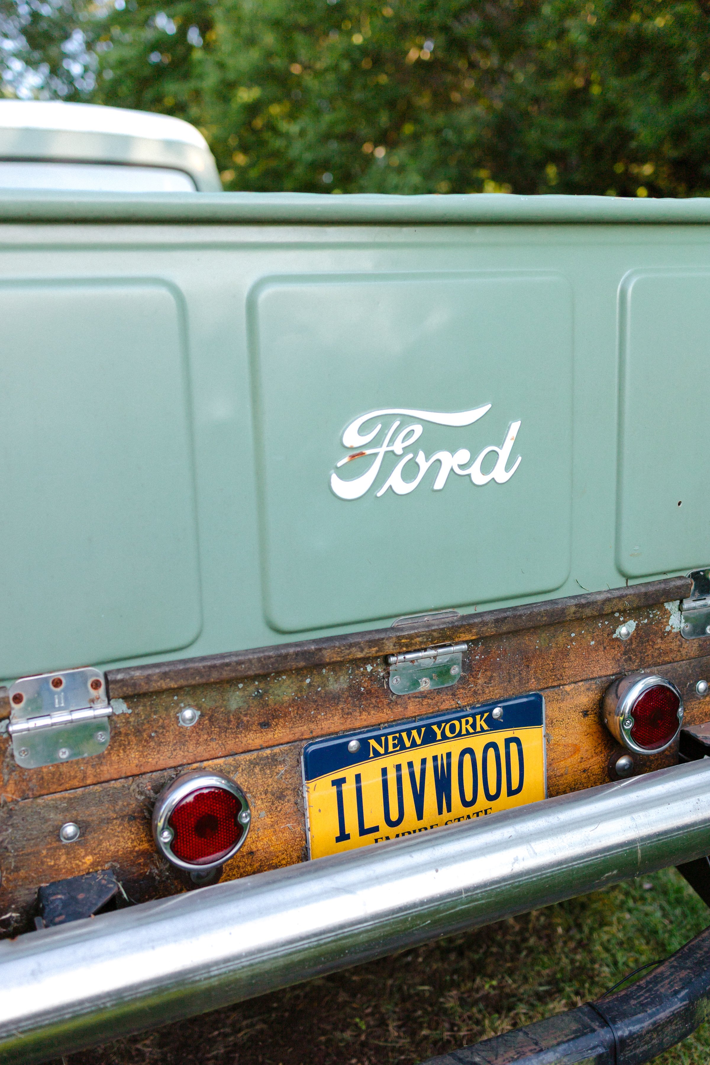 Close-up of the back of a vintage Ford truck with a green bed, New York license plate that reads 'ILUVWOOD,' and rusty wooden tailgate with two red taillights.