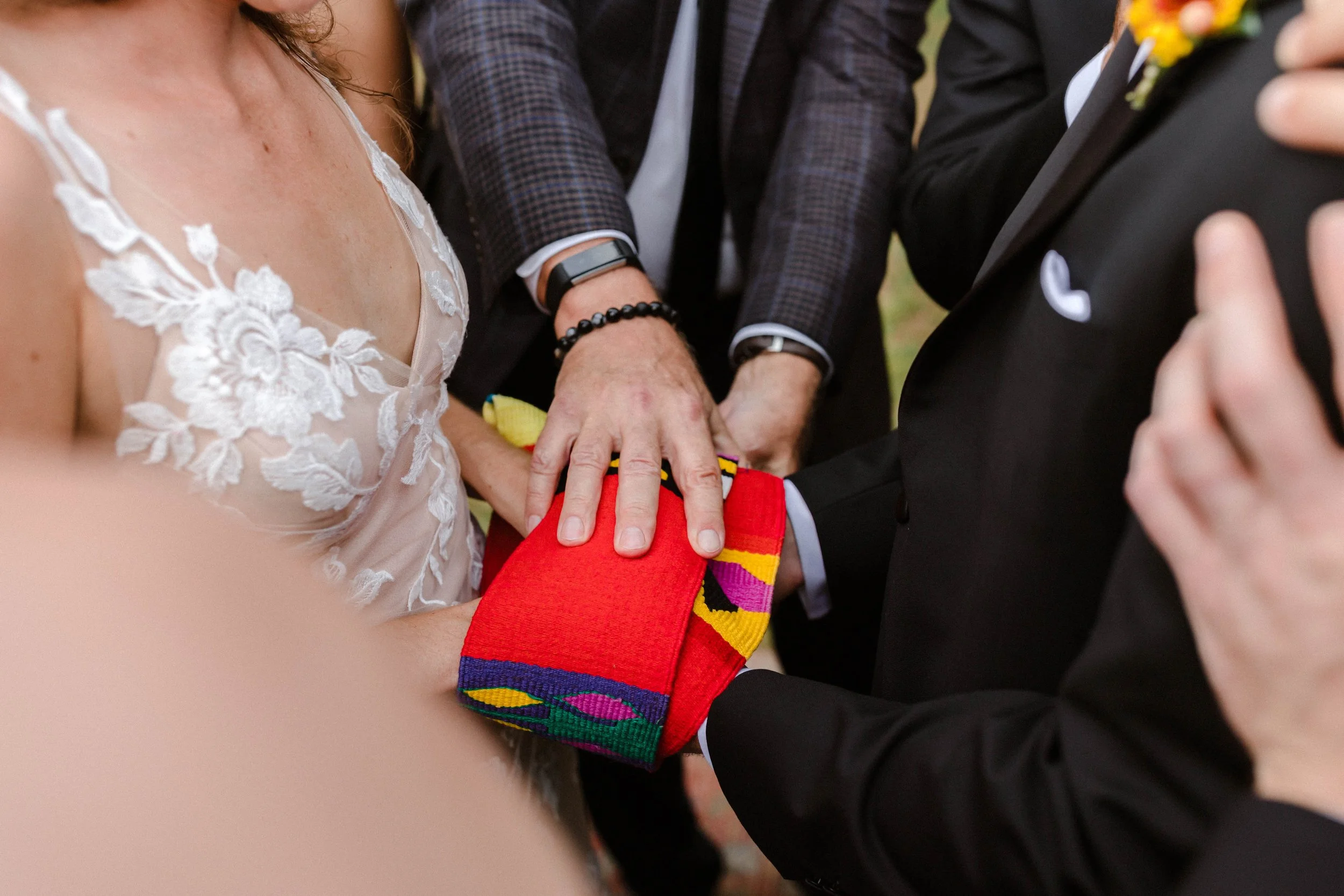 Close-up of a wedding ceremony showing a bride and groom with officiant's hand on top of a red and colorful patterned cloth on the couple's hands.