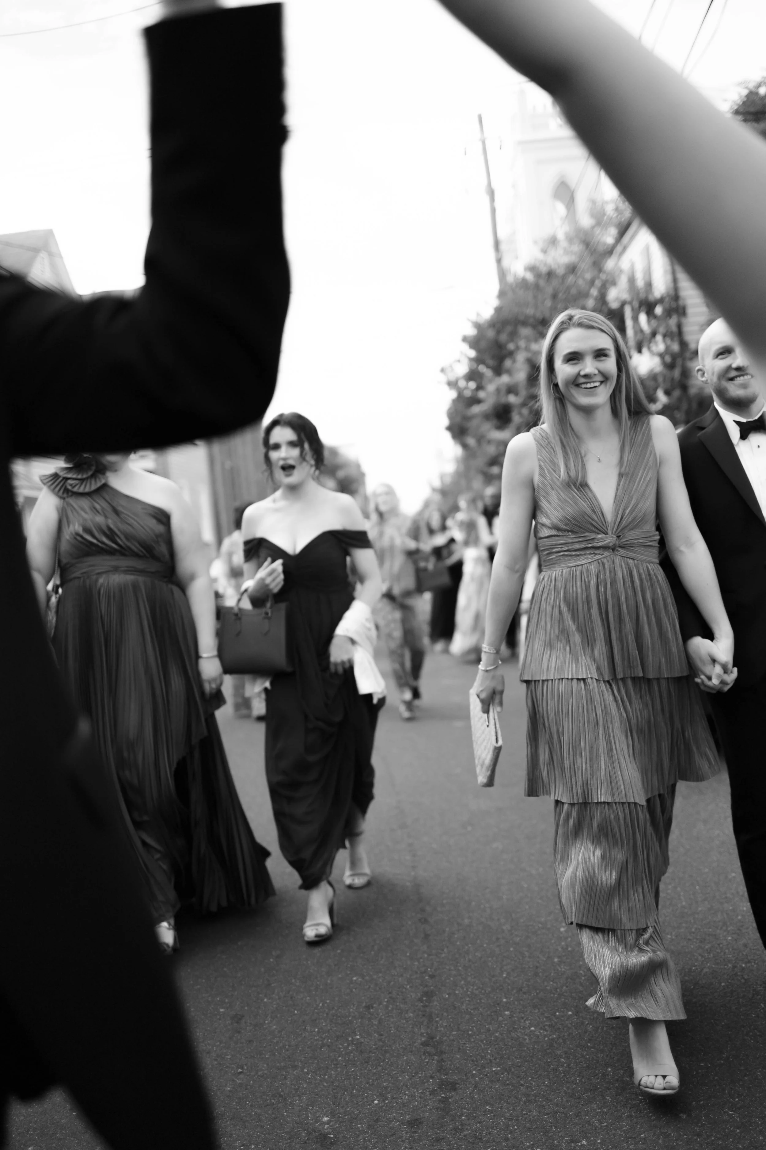 People dressed in formal attire walking on a street, smiling, and holding hands, in an outdoor event or celebration.
