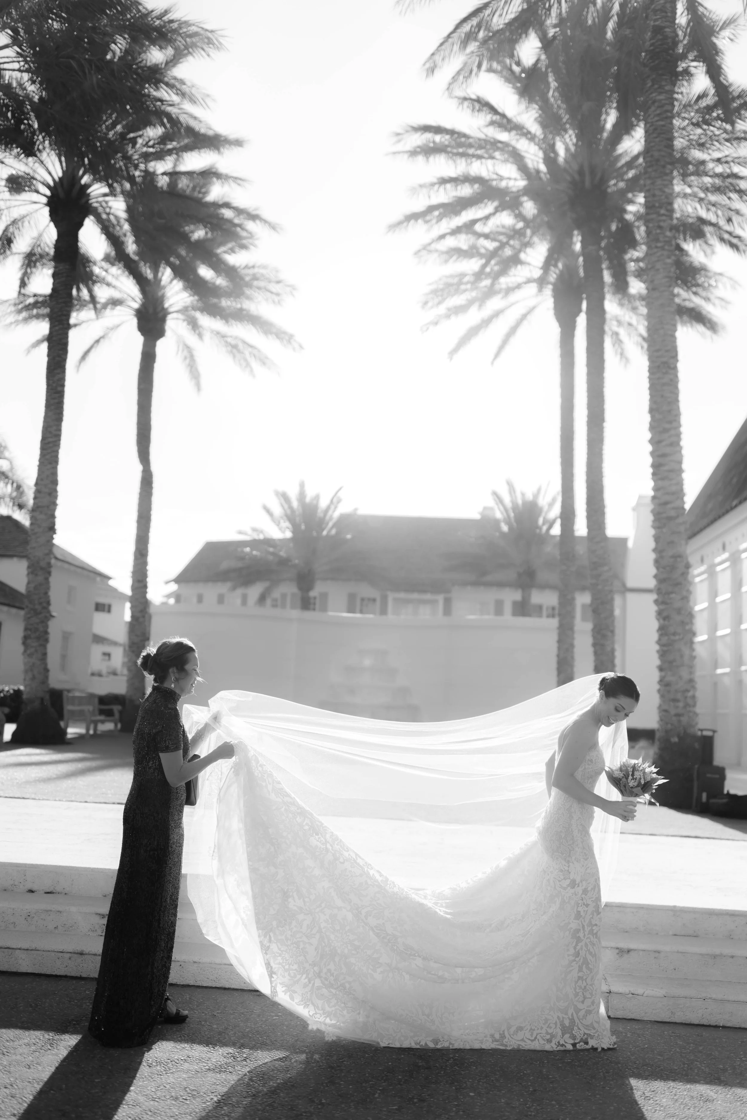A bride in a wedding dress holding a bouquet, with a woman holding her veil behind her, standing outdoors with palm trees and buildings in the background.