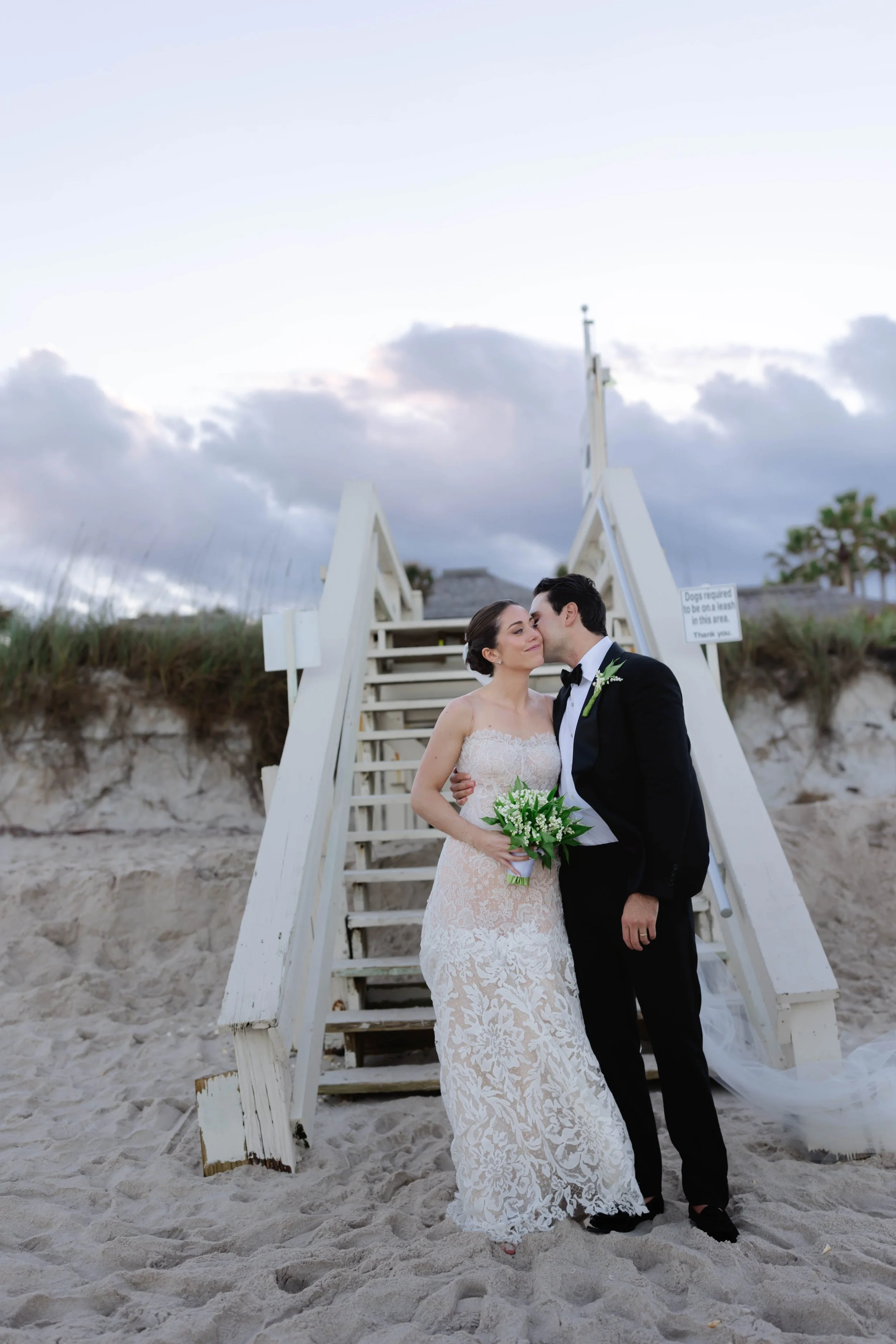 Bride and groom sharing a kiss on a sandy beach, standing in front of a white staircase, with cloudy sky in the background.
