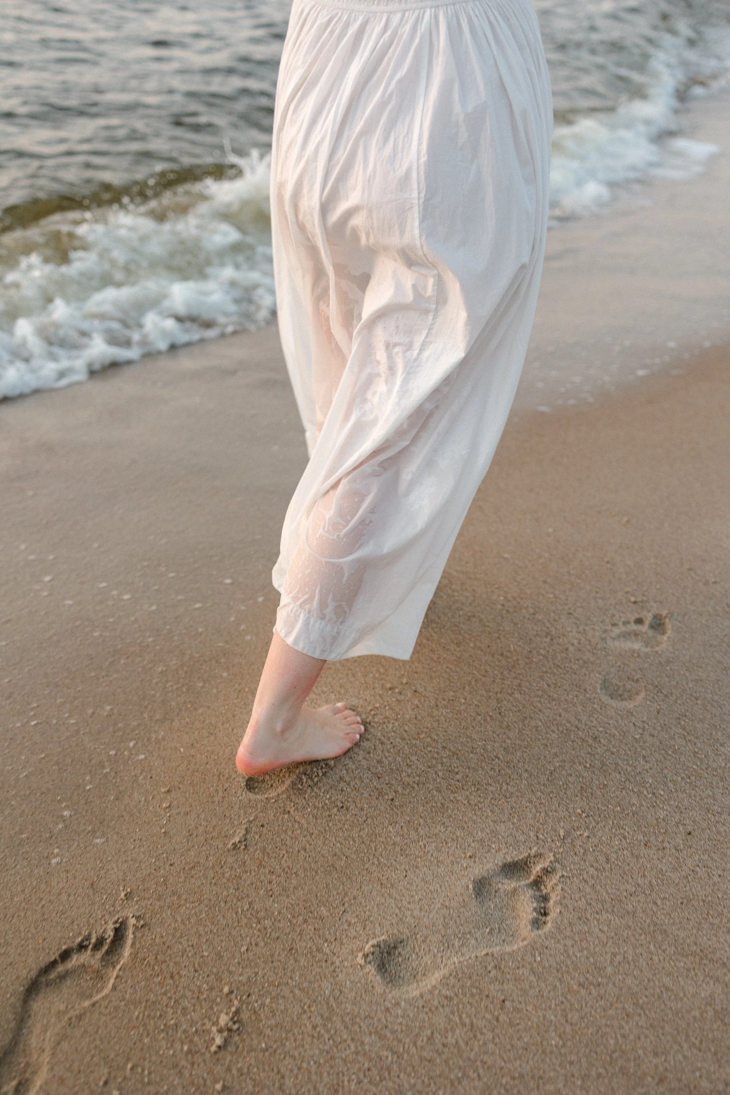 Person walking barefoot on sandy beach near the ocean, wearing white clothing.