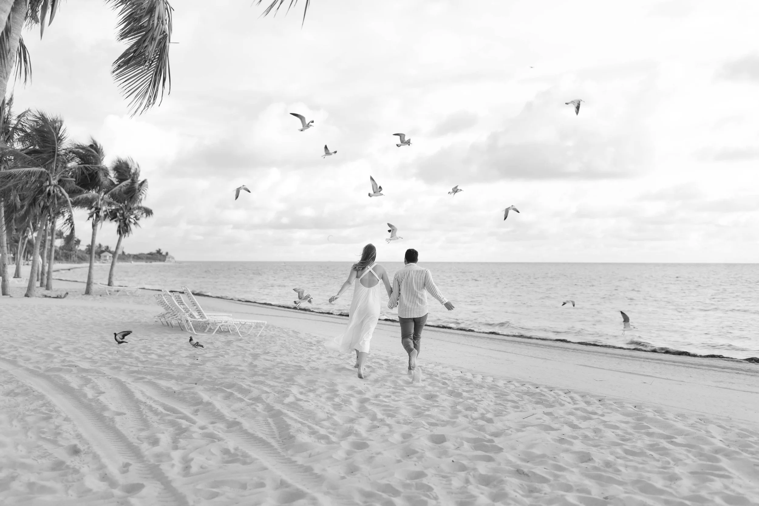 A couple holding hands while walking on the beach with palm trees on the left and seagulls flying overhead in the sky.