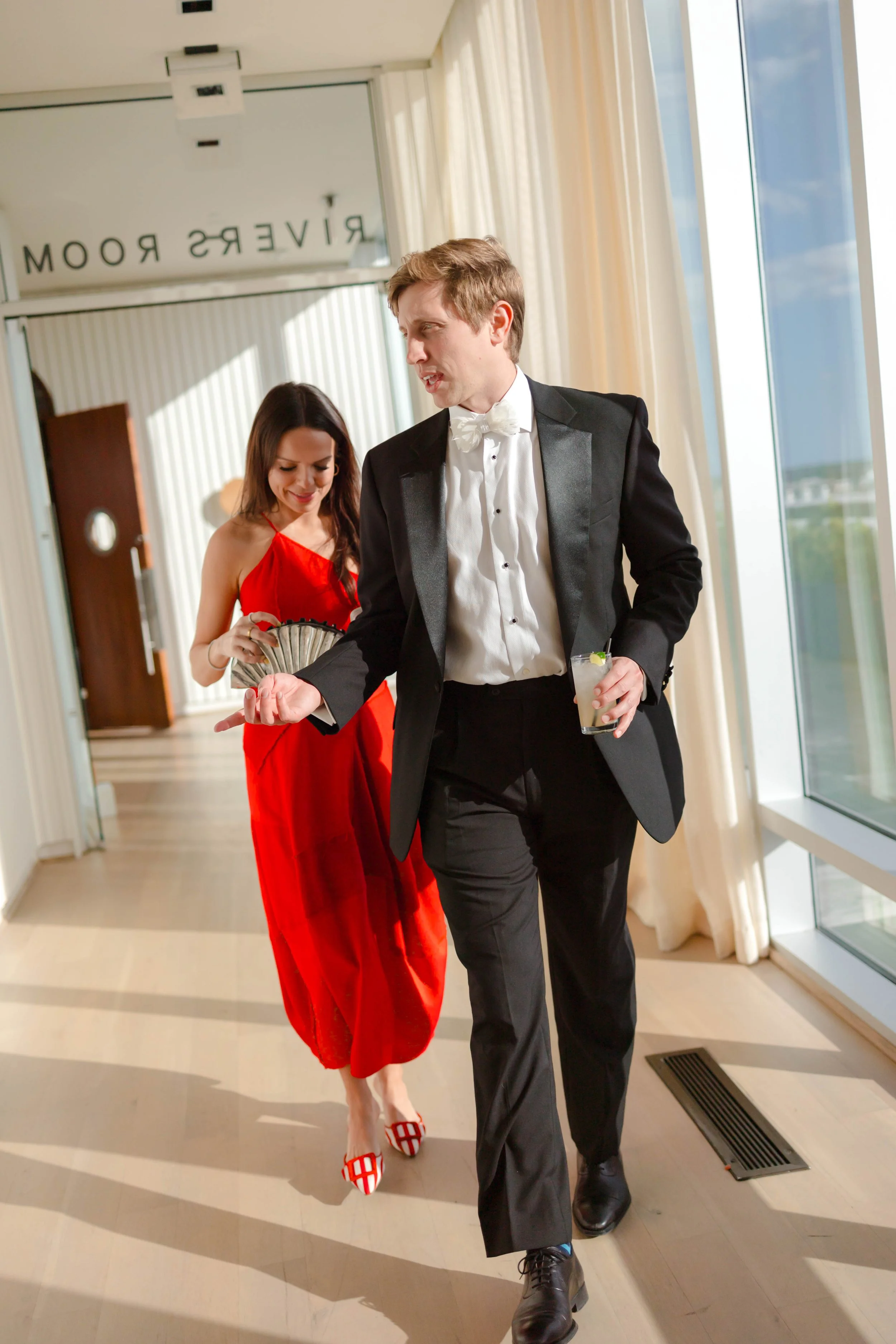 A man in a black tuxedo and a woman in a red dress walking through an elegant room with large windows, sunlight, and a sign that reads 'RIVERS ROOM'. The man holds a drink, and the woman holds a fan.