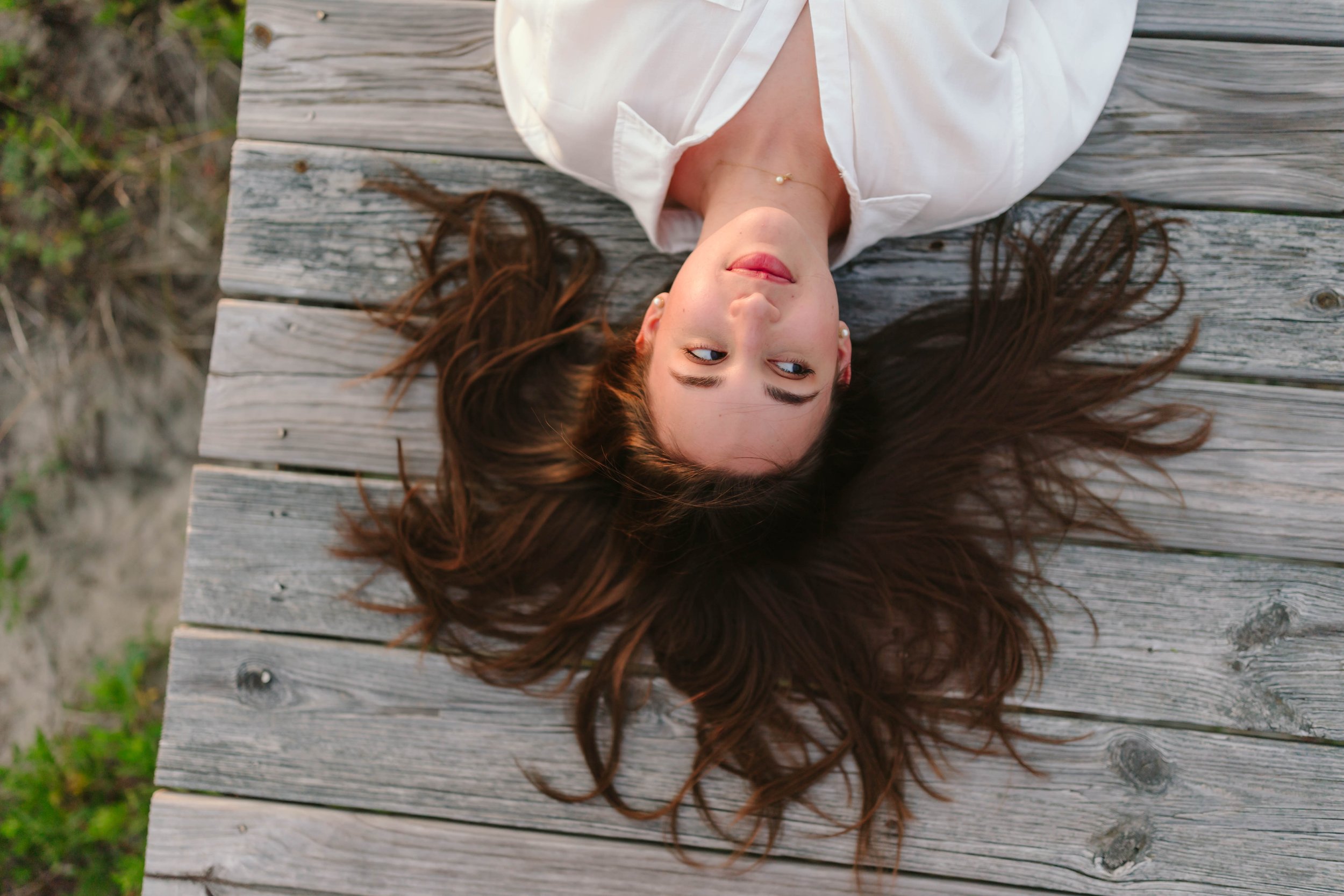 A young woman with long brown hair lies on her back on a wooden dock, wearing a white shirt, looking to the side.