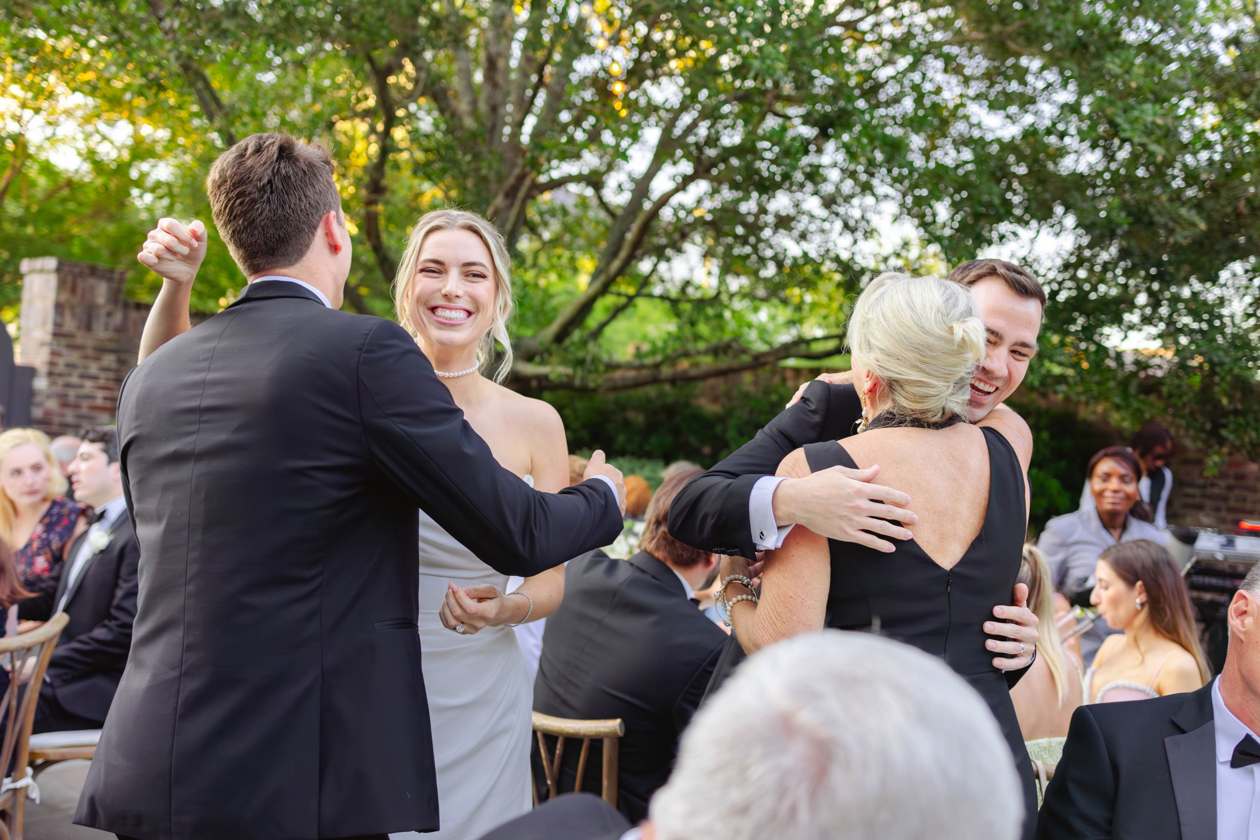 People dancing and hugging at an outdoor wedding reception with green trees in the background.
