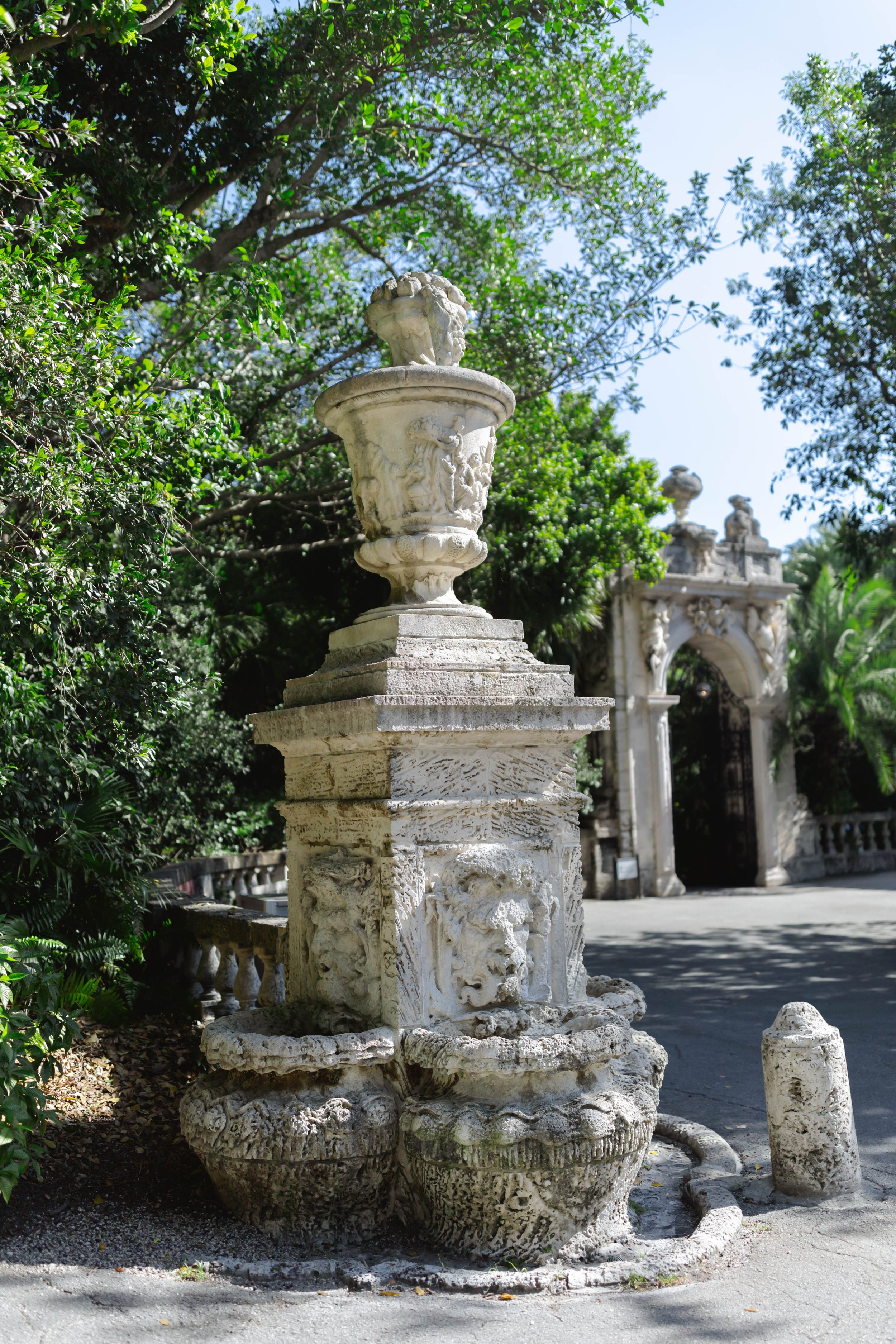 Ancient stone fountain with carved lion heads in front of a historic archway, surrounded by green trees and bushes.