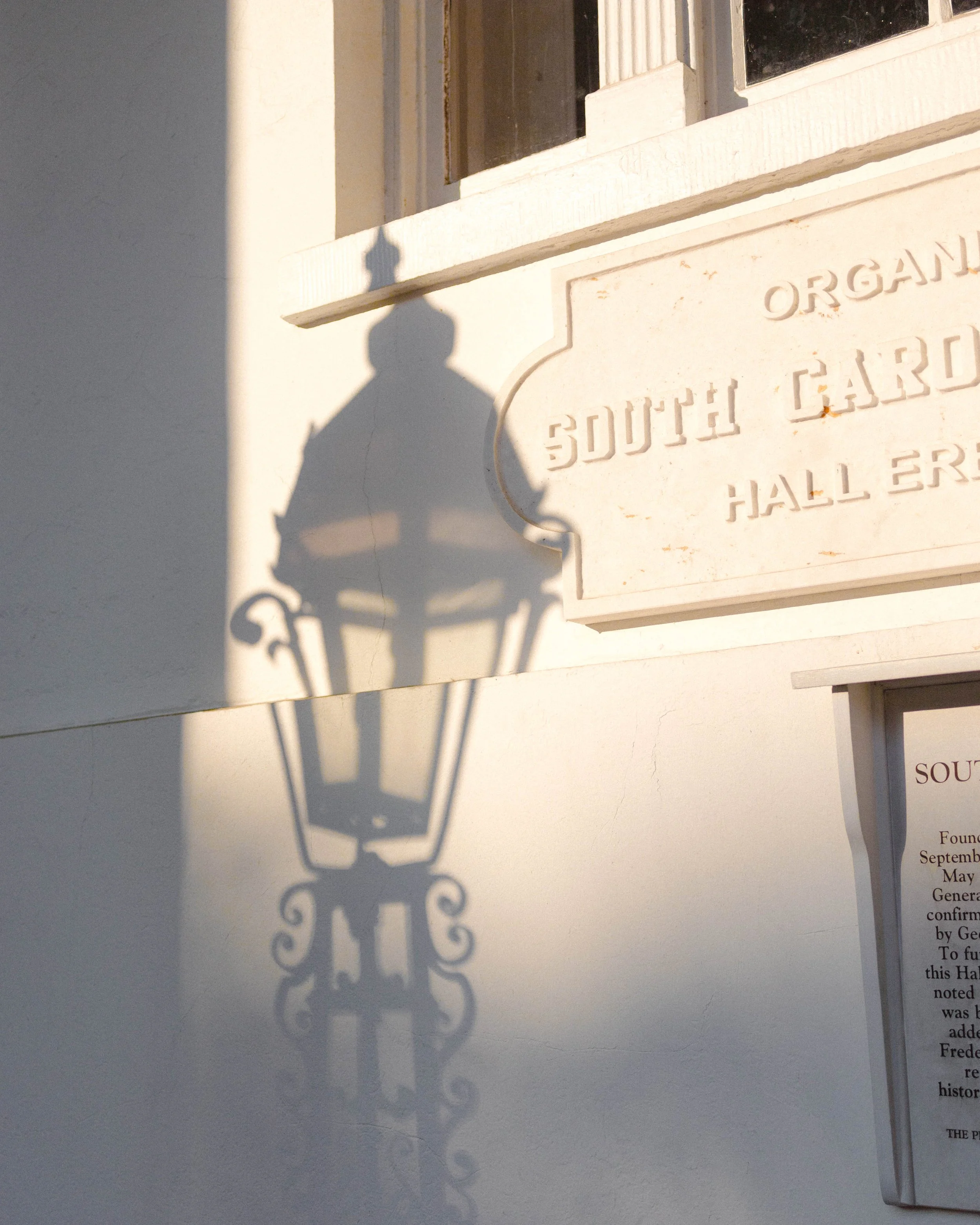 Shadow of a street lamp cast on a white wall with a plaque reading 'South Garden Hall' nearby.