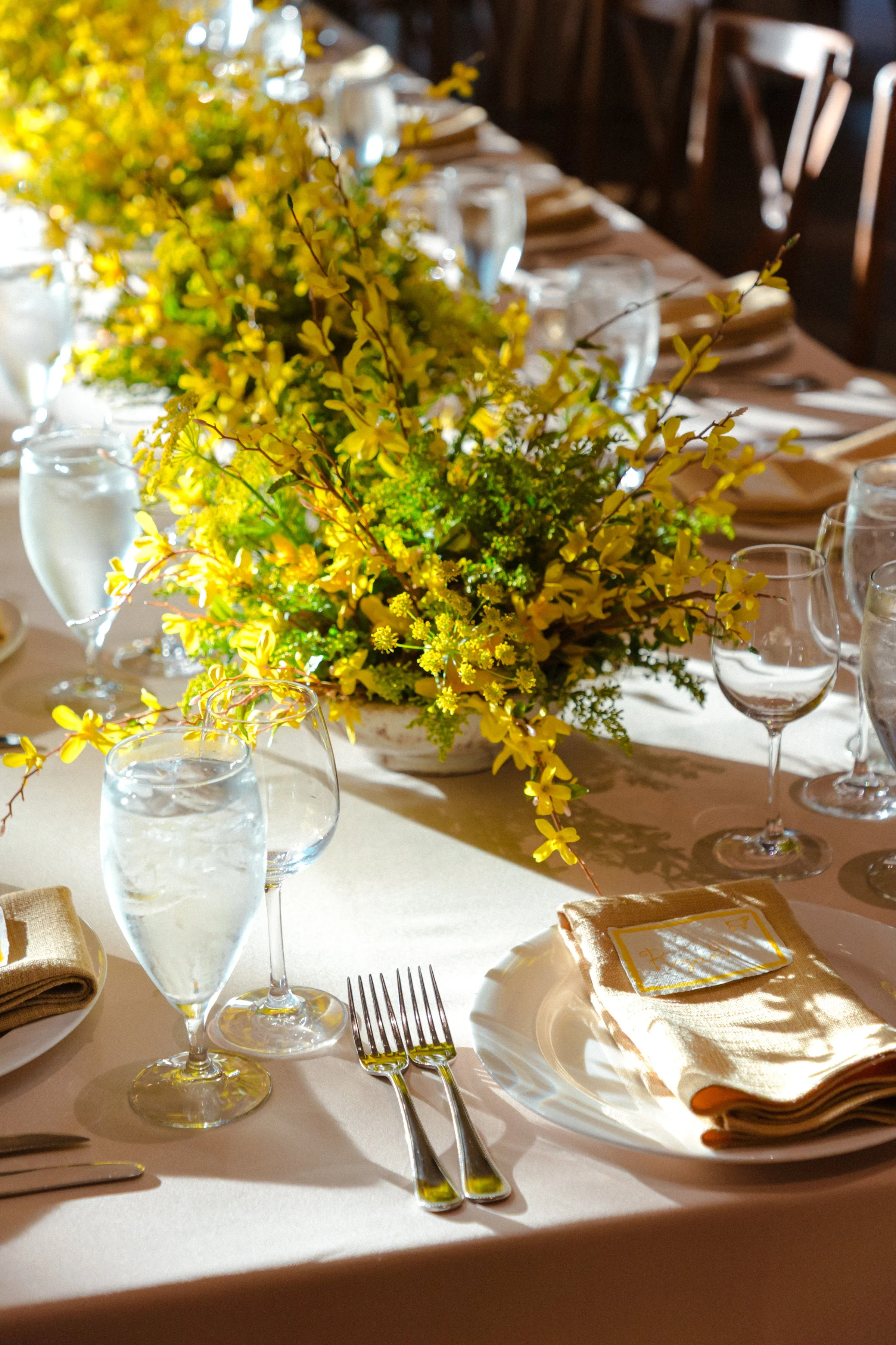 A table set for a meal with a floral centerpiece consisting of yellow flowers and greenery, water glasses, napkins, and silverware arranged for a formal event.