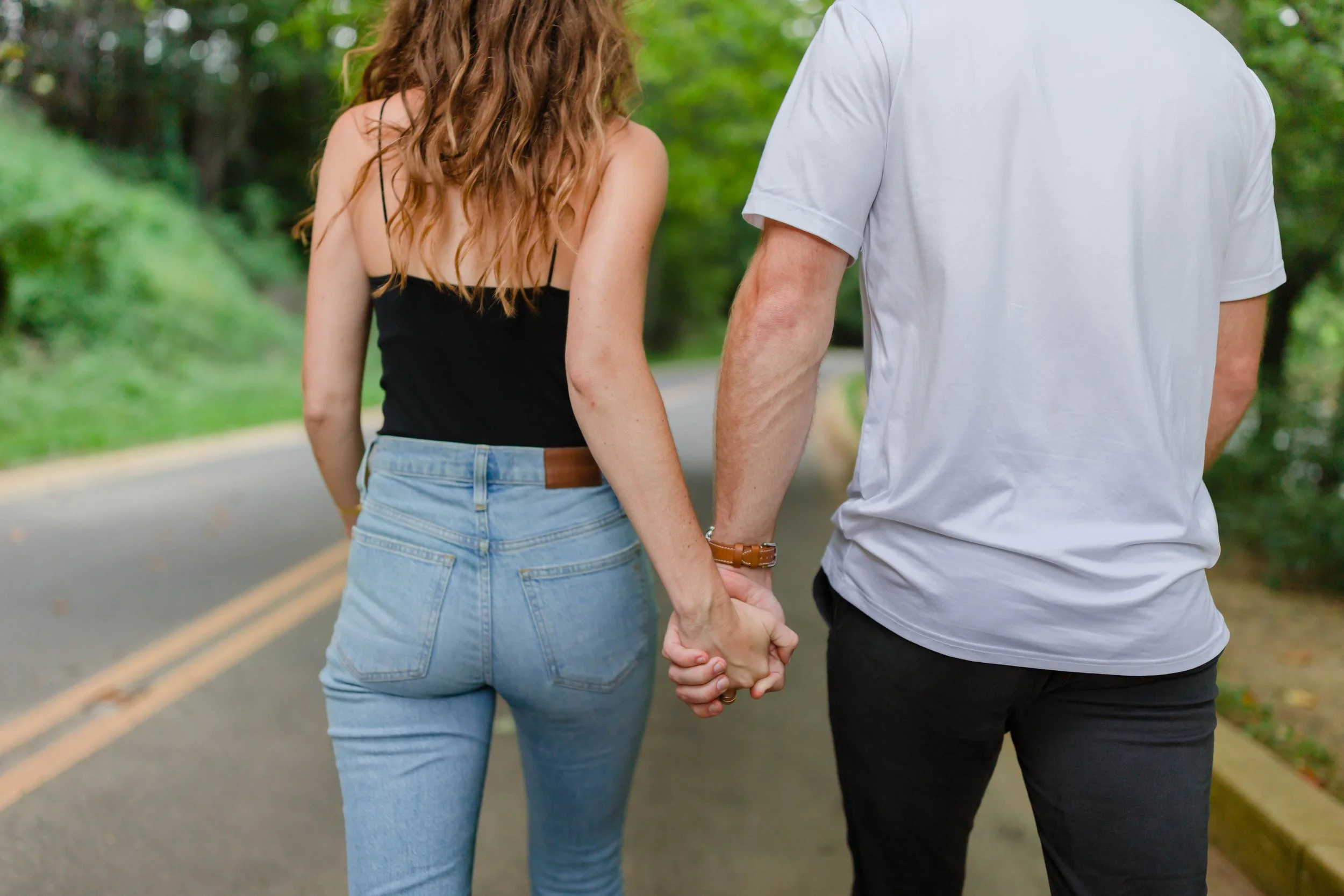 A couple walking hand in hand on a paved road surrounded by lush greenery.