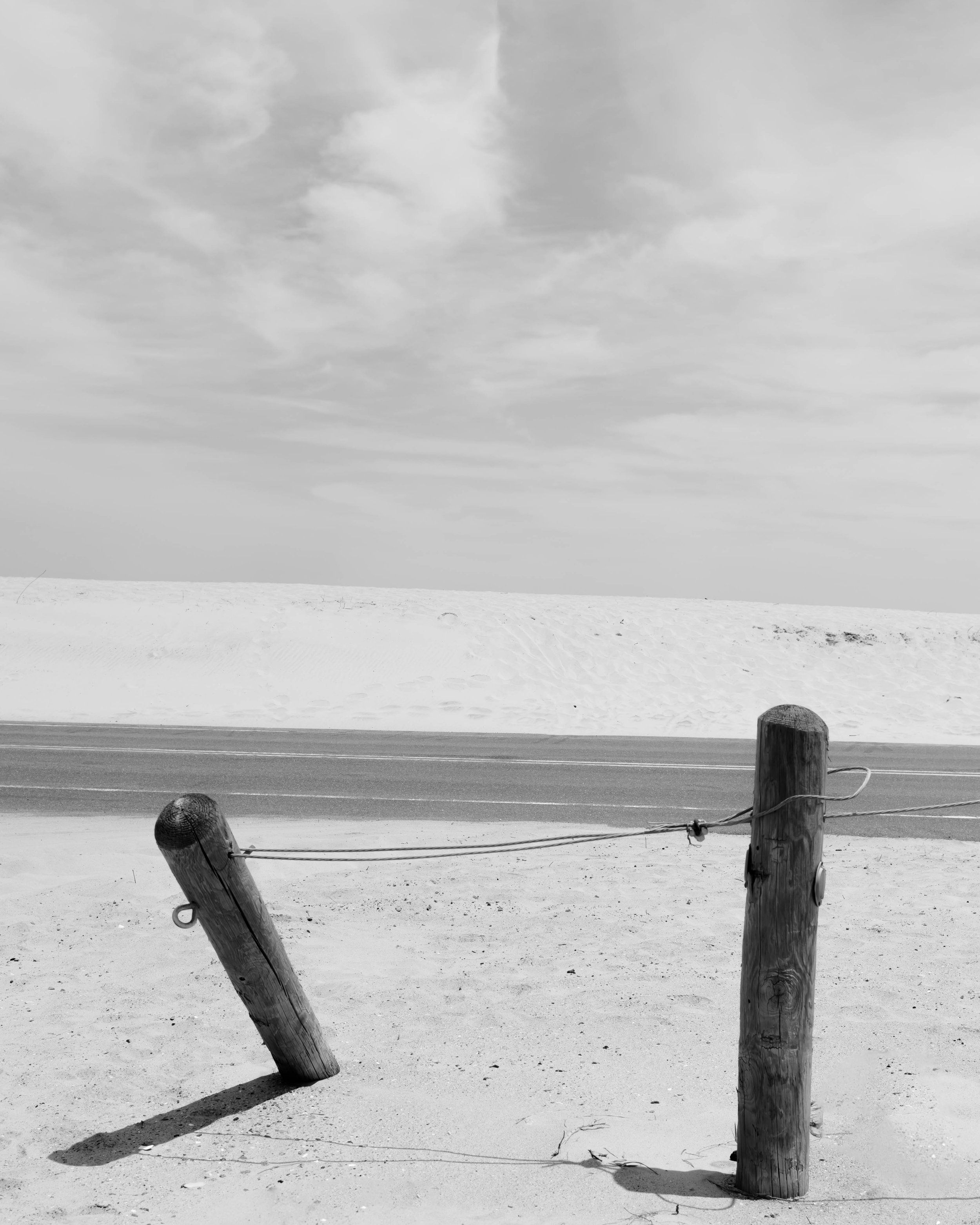 Black and white photograph of a sandy beach with two weathered wooden posts connected by a rope, with dunes and a cloudy sky in the background.