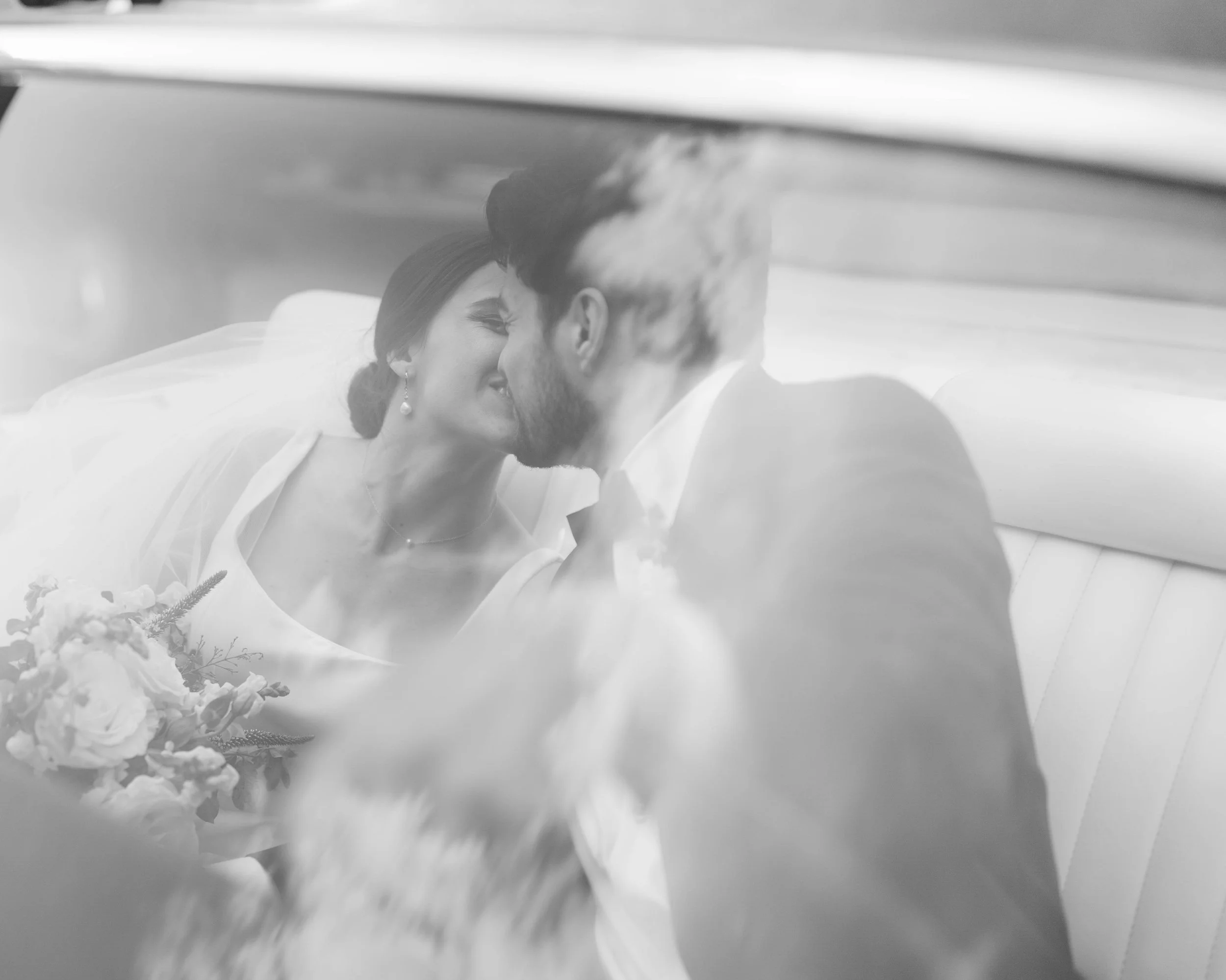 A bride and groom sitting inside a vehicle, sharing a kiss after their wedding ceremony, with the bride holding a bouquet of flowers.