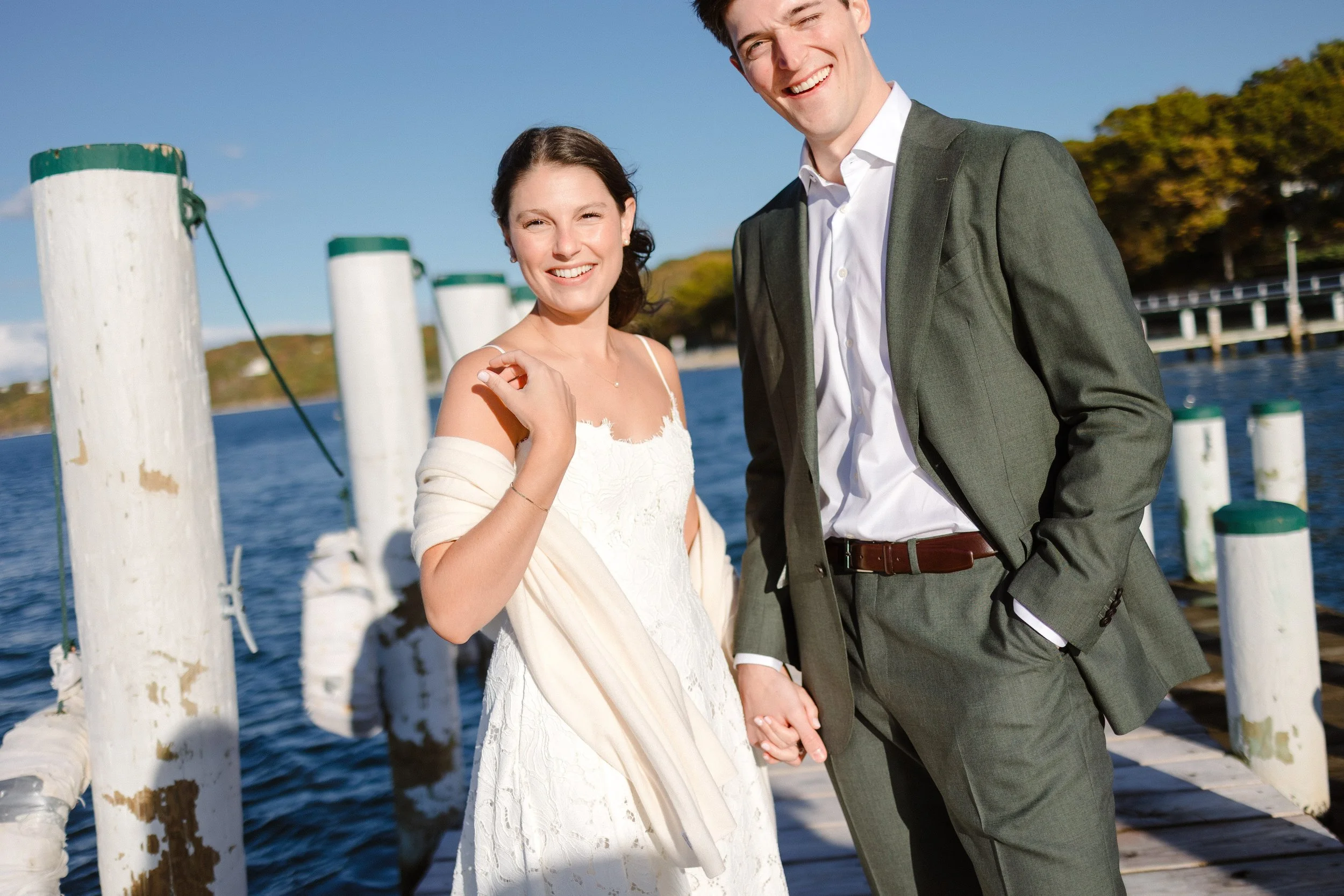 A smiling couple holding hands on a dock by the water, dressed in formal attire, with the woman in a white dress and the man in a gray suit, under a blue sky and trees in the background.
