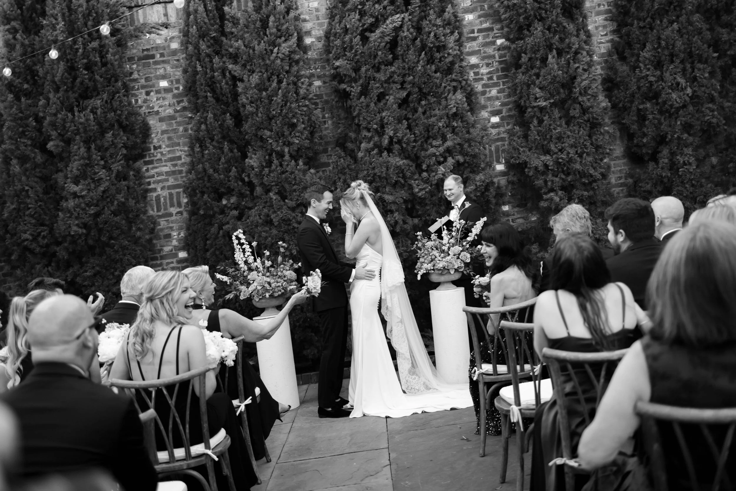 A black and white photo of a wedding ceremony with the bride and groom in the center, smiling and holding hands, surrounded by seated guests. The bride is in a white dress with a veil, and the groom is in a dark suit. There are large floral arrangeme