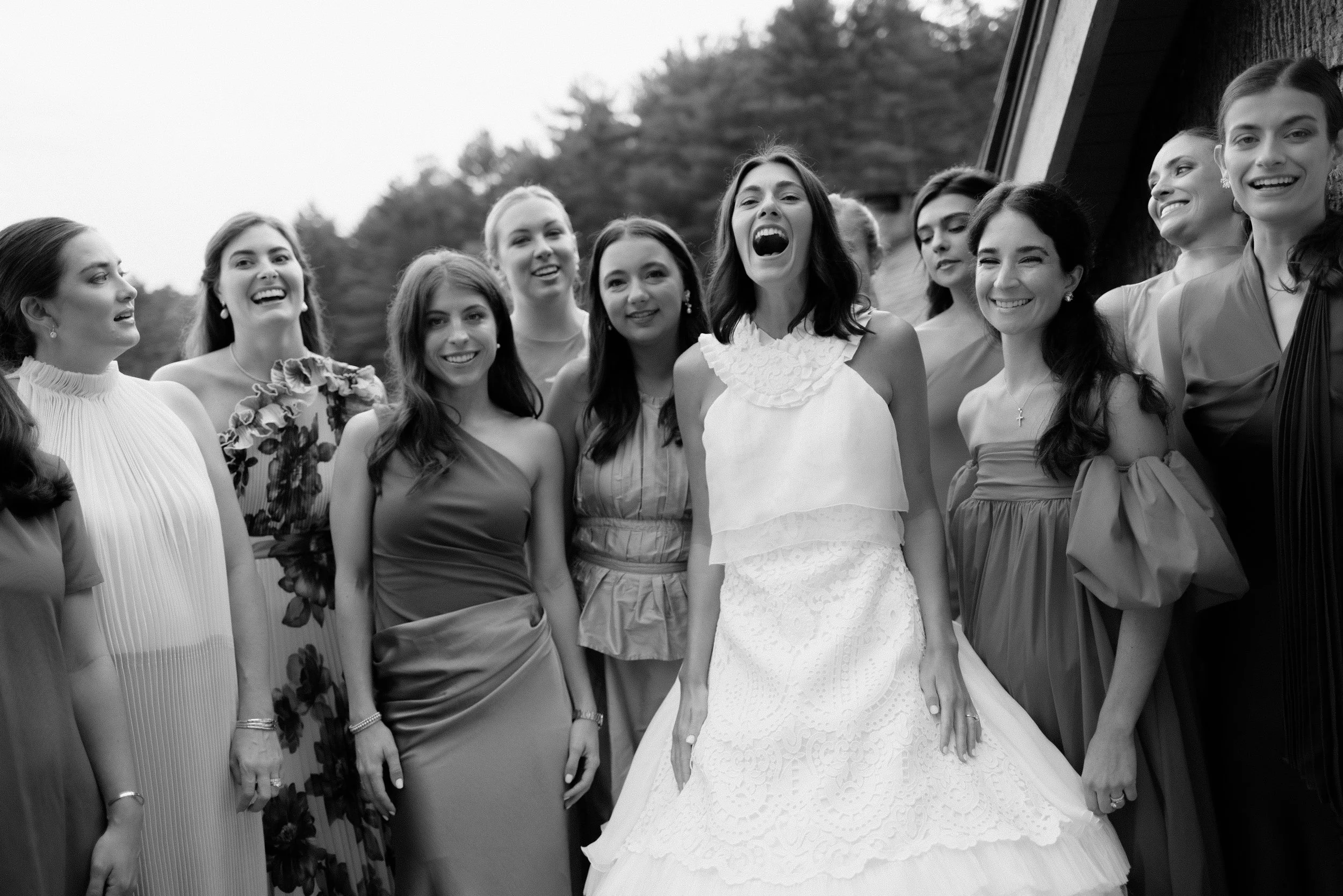 A group of women at a wedding, with the bride in the center wearing a white gown, surrounded by bridesmaids in various dresses, outdoors with trees in the background.