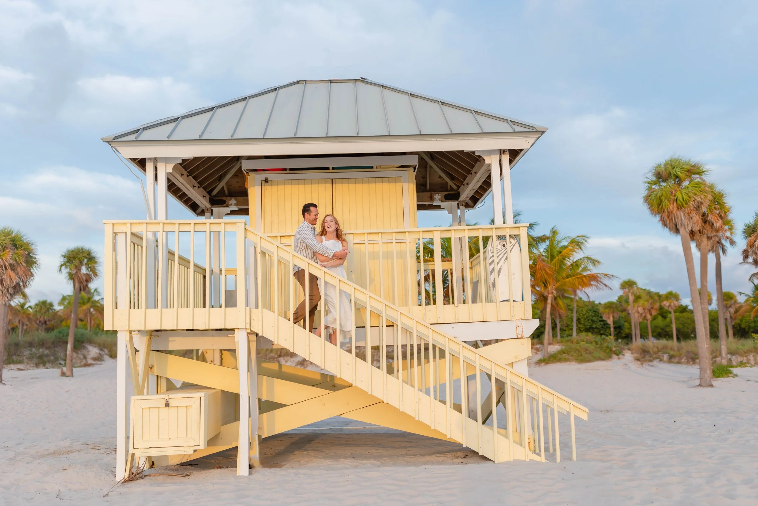 A couple dancing on a yellow lifeguard tower on a sandy beach with palm trees in the background.