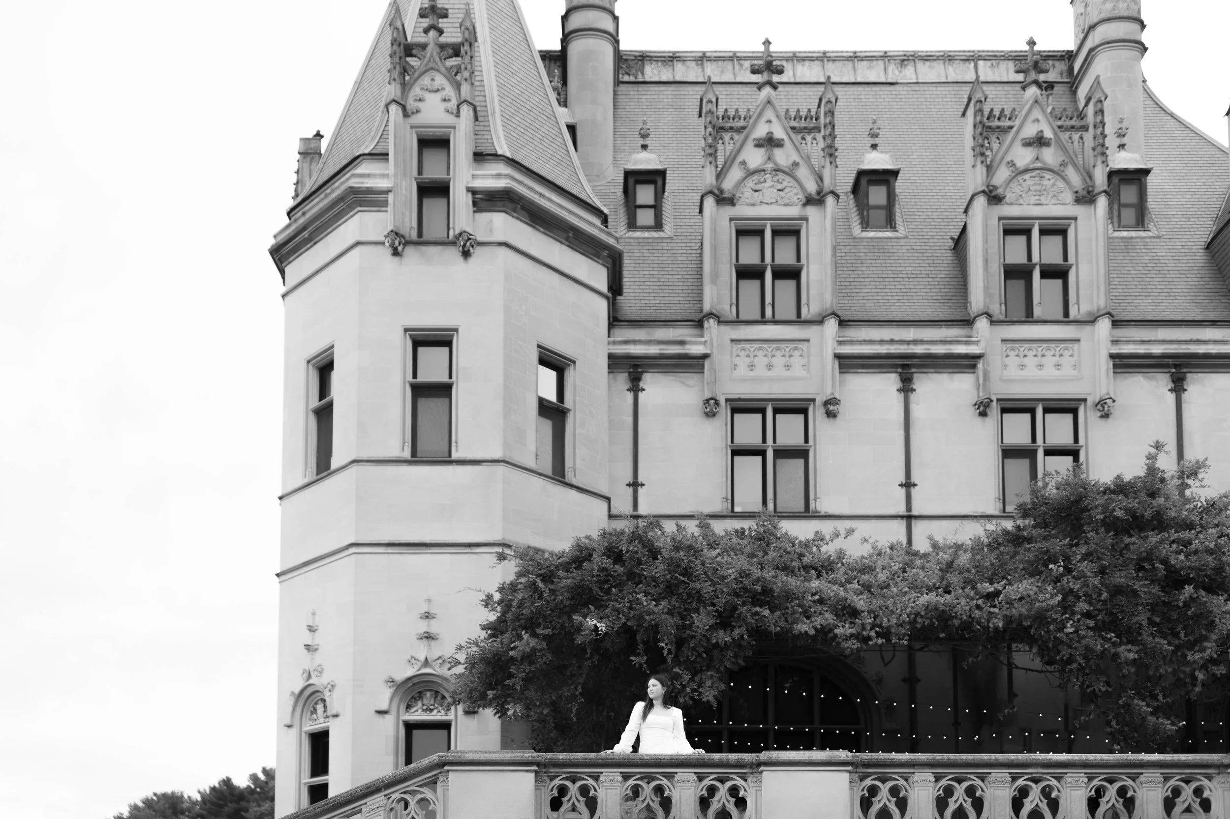 Black and white photo of a woman standing on a castle-like building's balcony with ornate stone railing, large tower, multiple windows, and decorative architectural details.