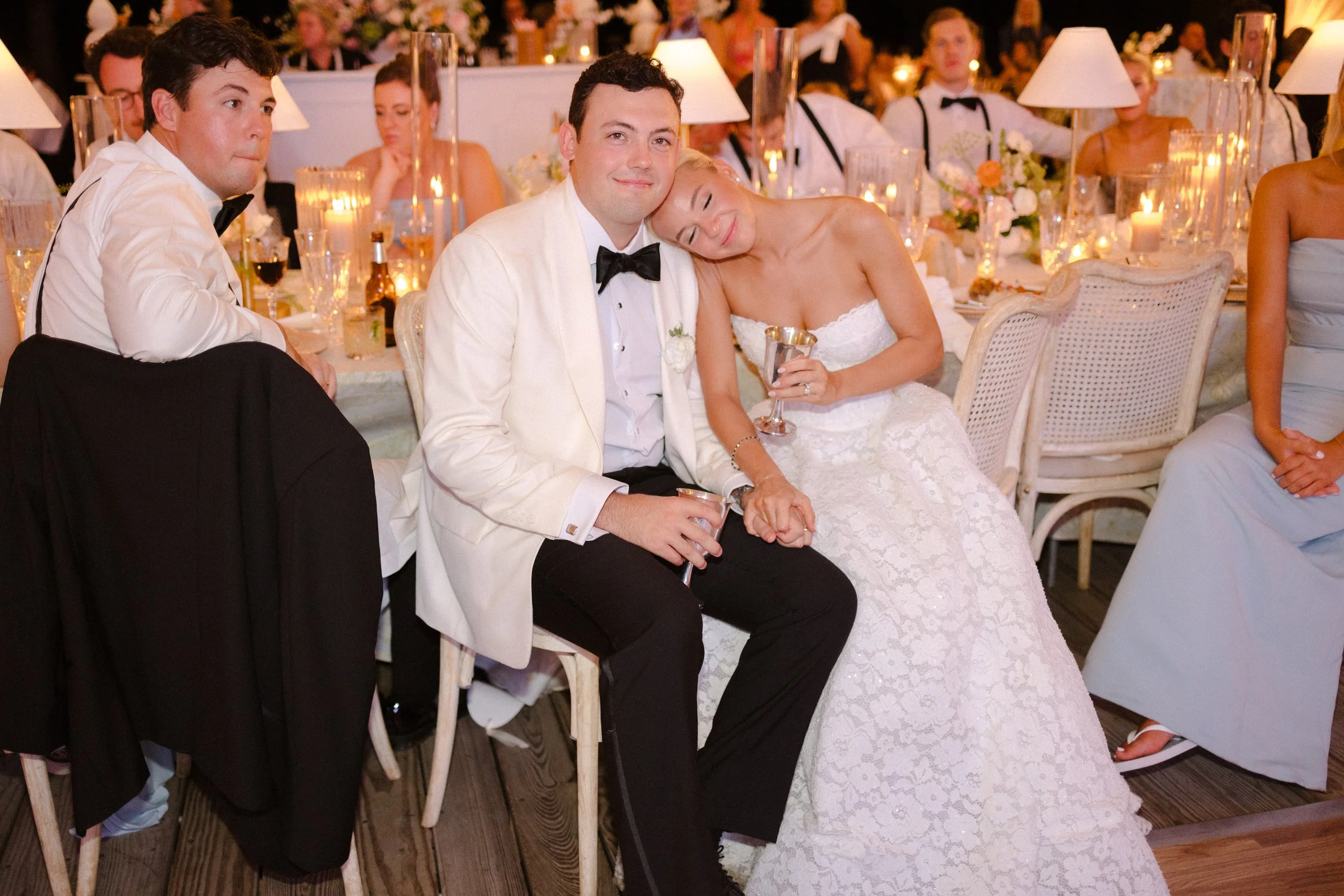 A bride and groom sitting together at a wedding reception, holding hands and smiling. The bride is resting her head on the groom's shoulder, and they are surrounded by guests seated at tables with candles and flowers.