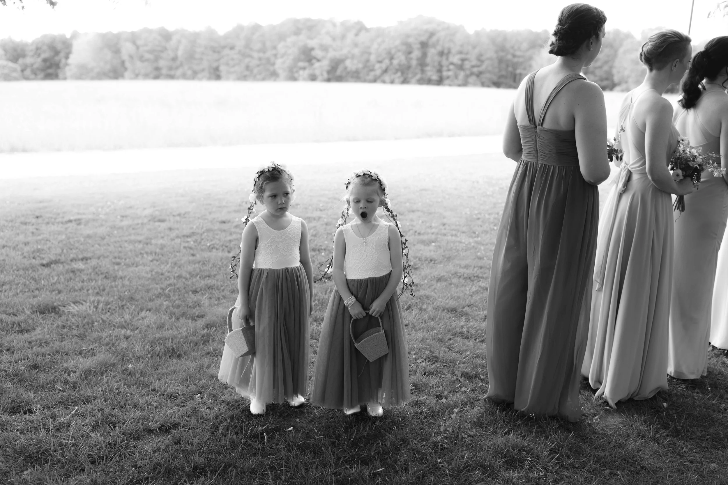Two young girls in formal dresses standing on grass, one yawning, with women dressed in formal attire nearby, outside on a sunny day.