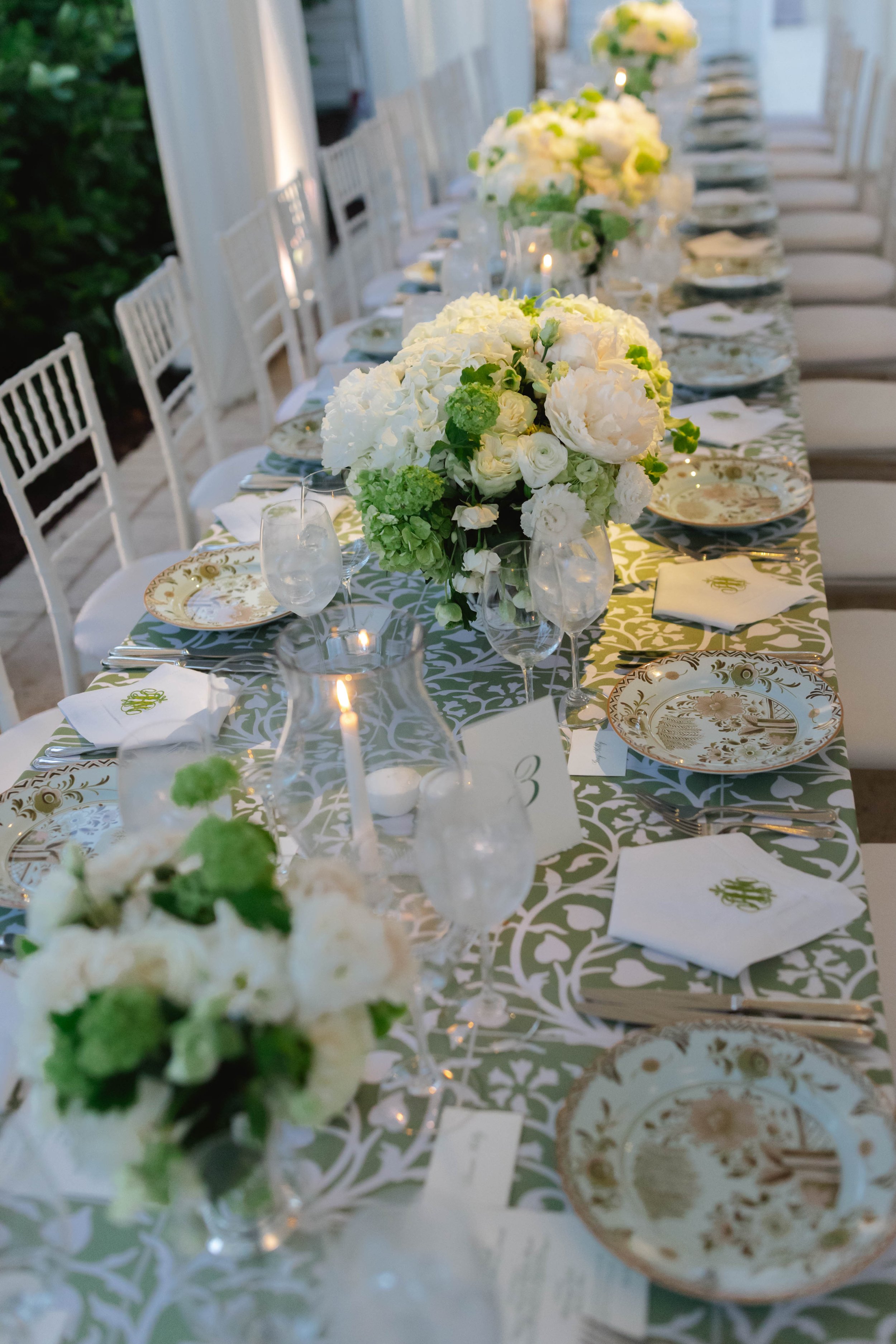 Long banquet table set with white floral centerpieces, gold and white patterned plates, glasses, and napkins with monogrammed initials, decorated for a formal event or wedding reception.