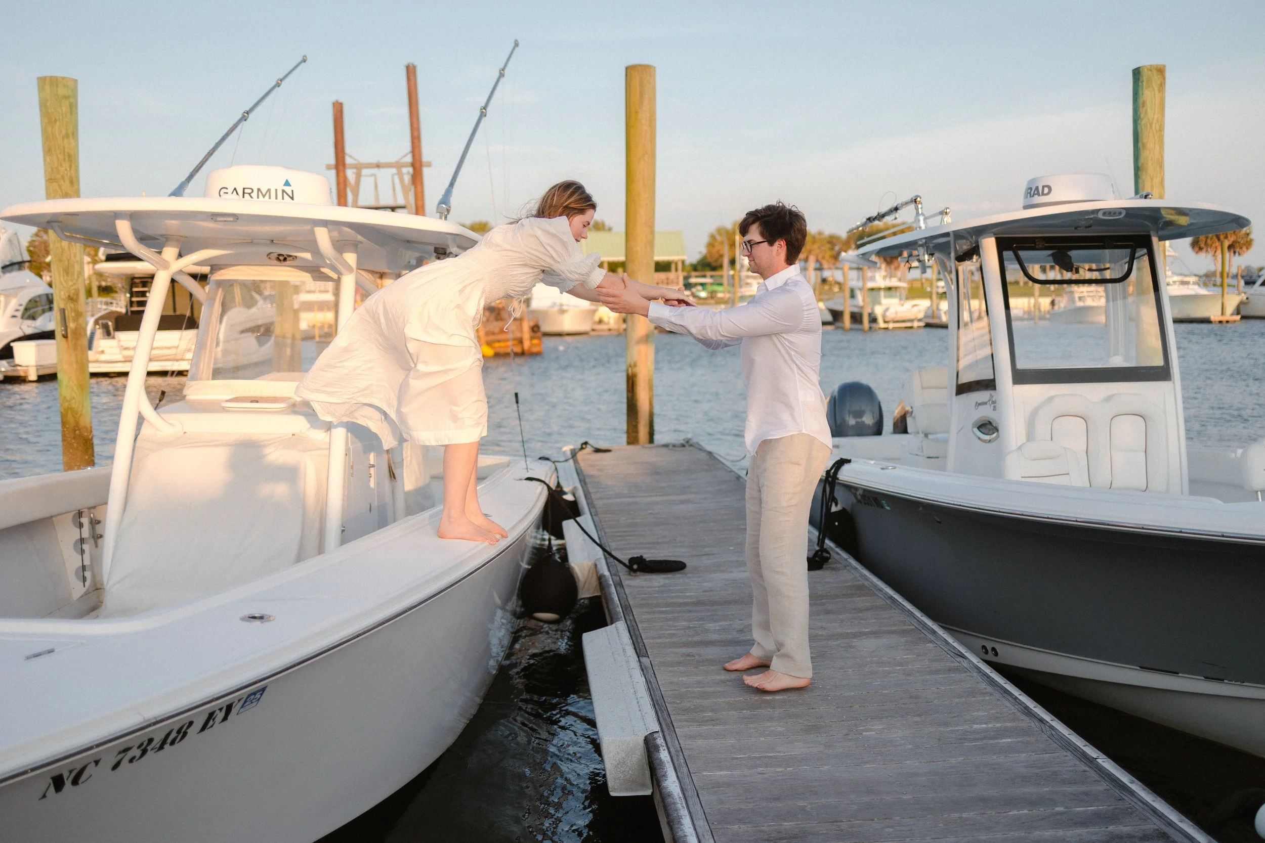 A woman helping a man aboard a boat at a marina with other boats docked in the background, during sunset.
