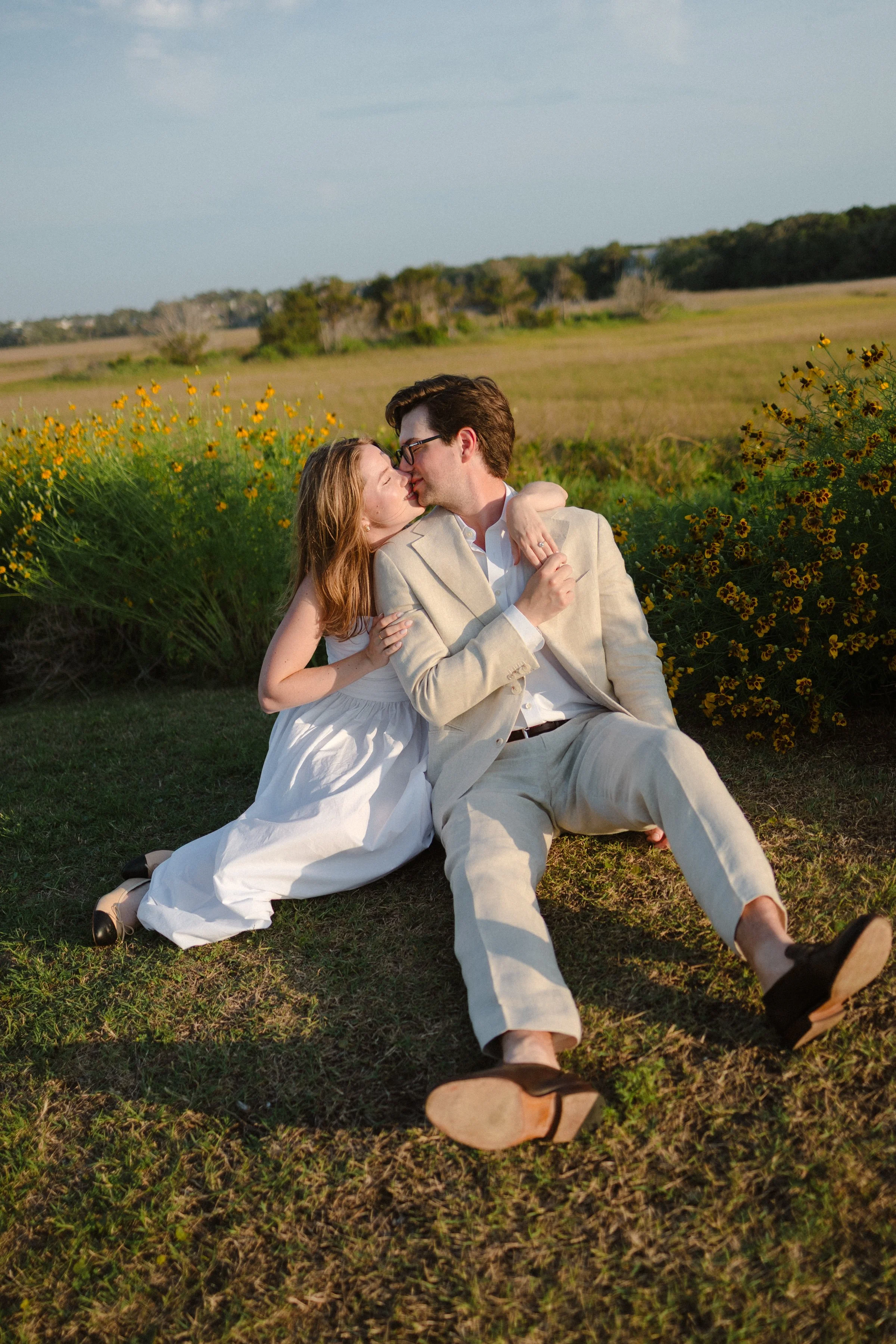 A couple sitting on grass in a field, leaning in for a kiss, with yellow flowers and open landscape in the background at sunset.