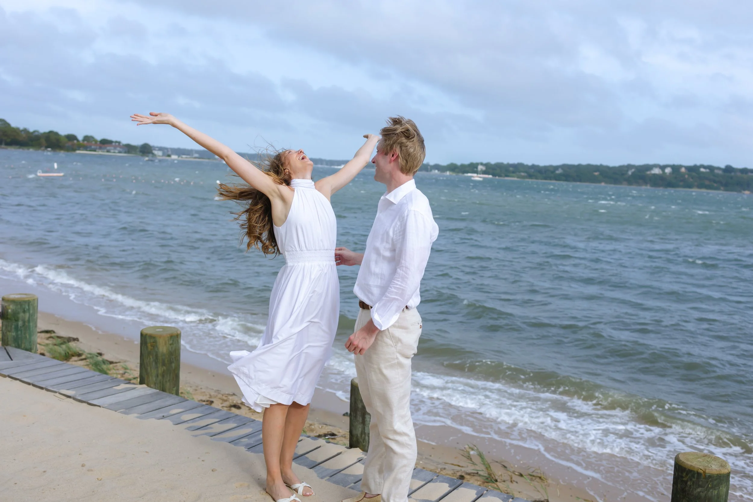 A joyful young woman and man dancing and laughing on a beach with water and a cloudy sky in the background.