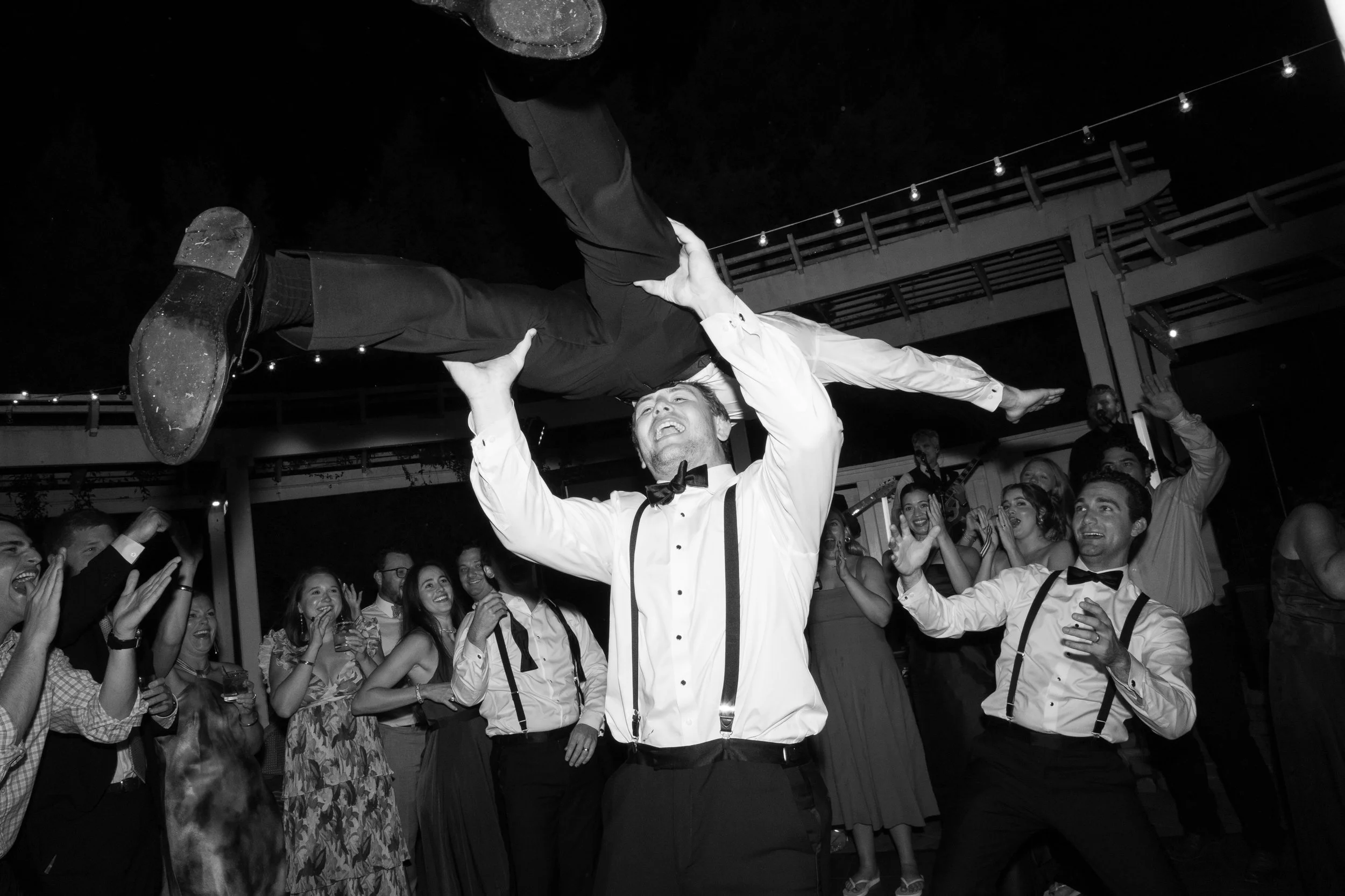 A man holding another man above his head at a party, with guests around celebrating and clapping. It is a black-and-white photo.