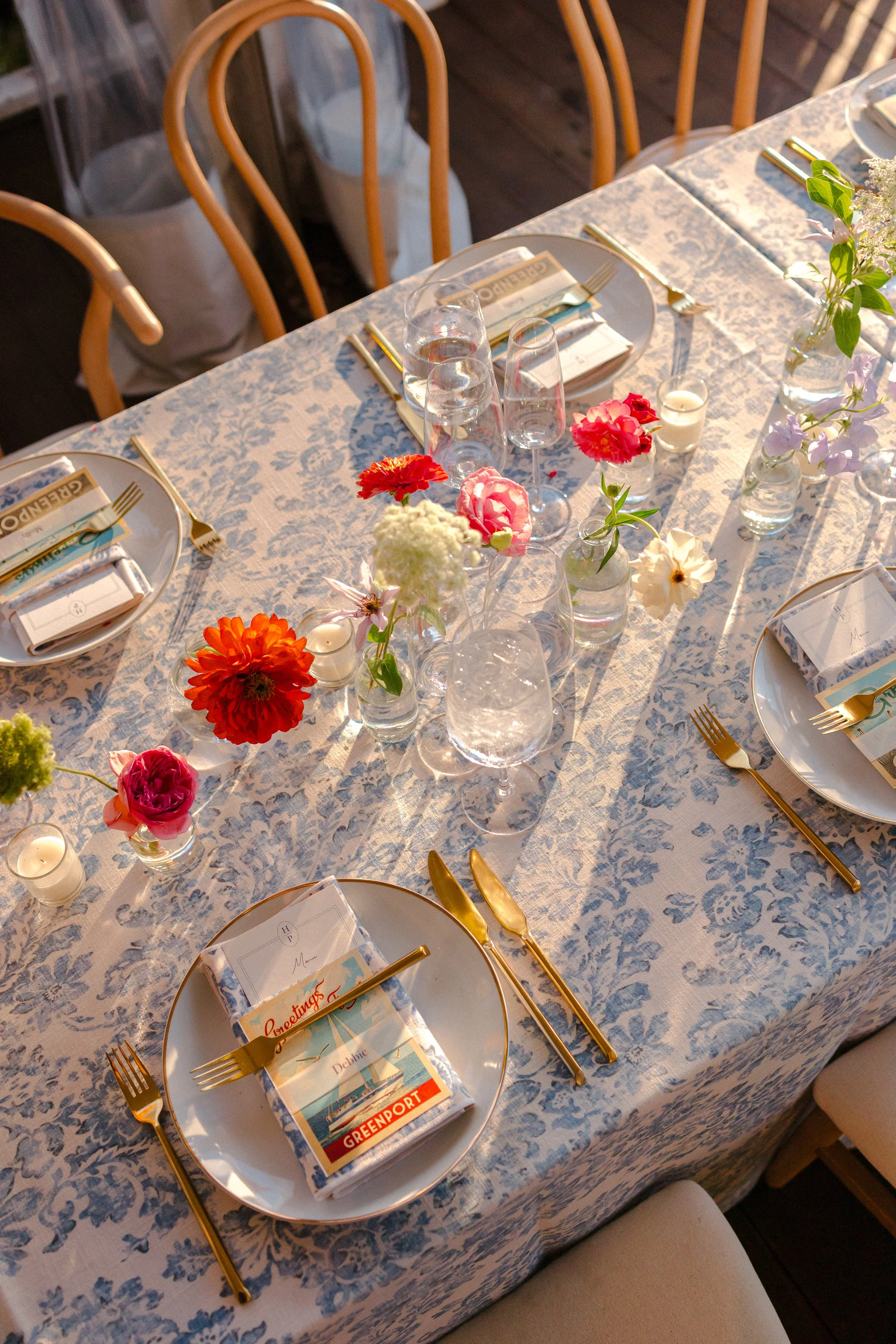 A dining table set for a meal with plates, gold utensils, floral arrangements, and glasses, covered with a patterned tablecloth.