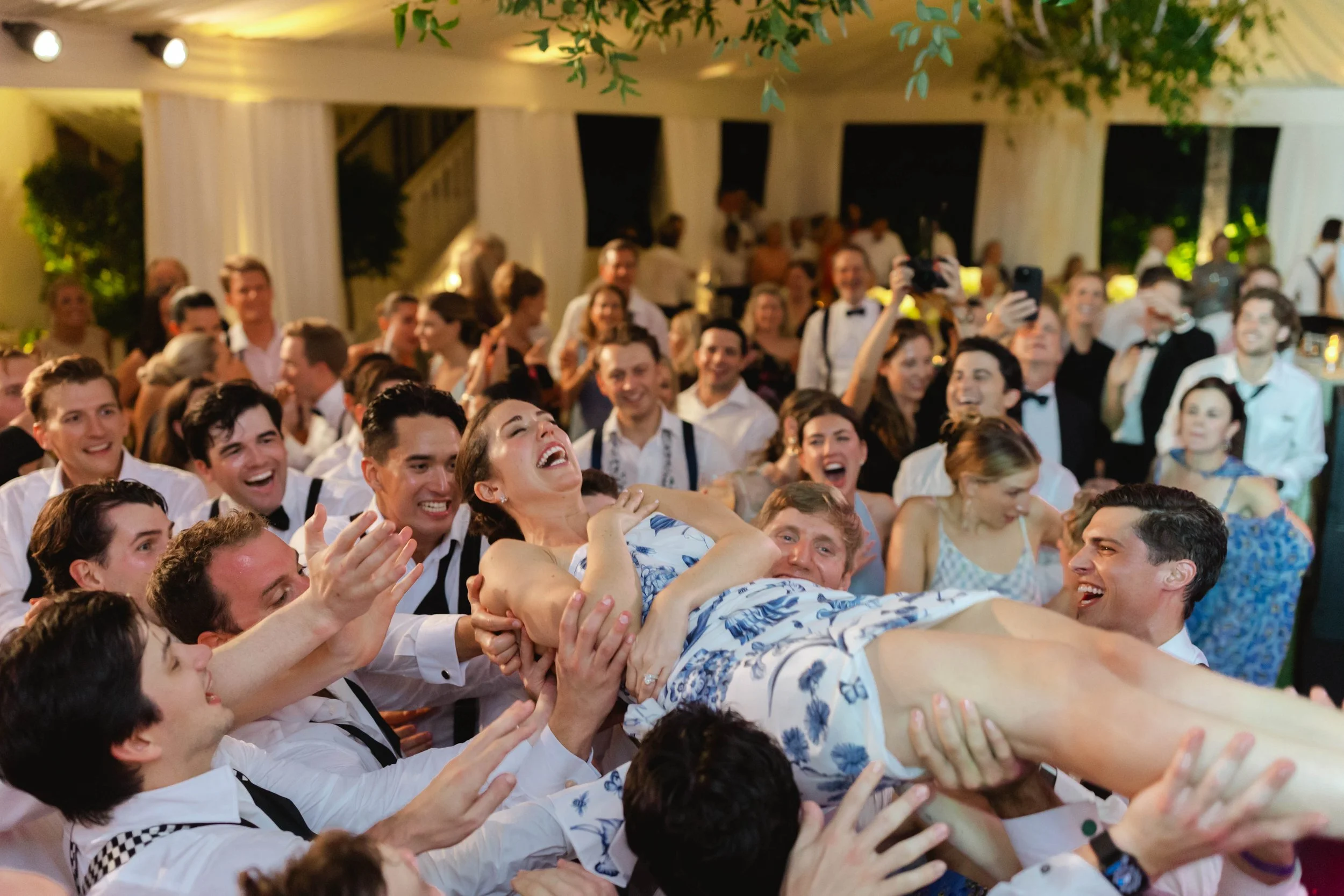 A large group of cheerful people at a wedding reception, lifting and passing a woman in a floral dress overhead, surrounded by friends in formal attire, inside an elegant decorated venue.