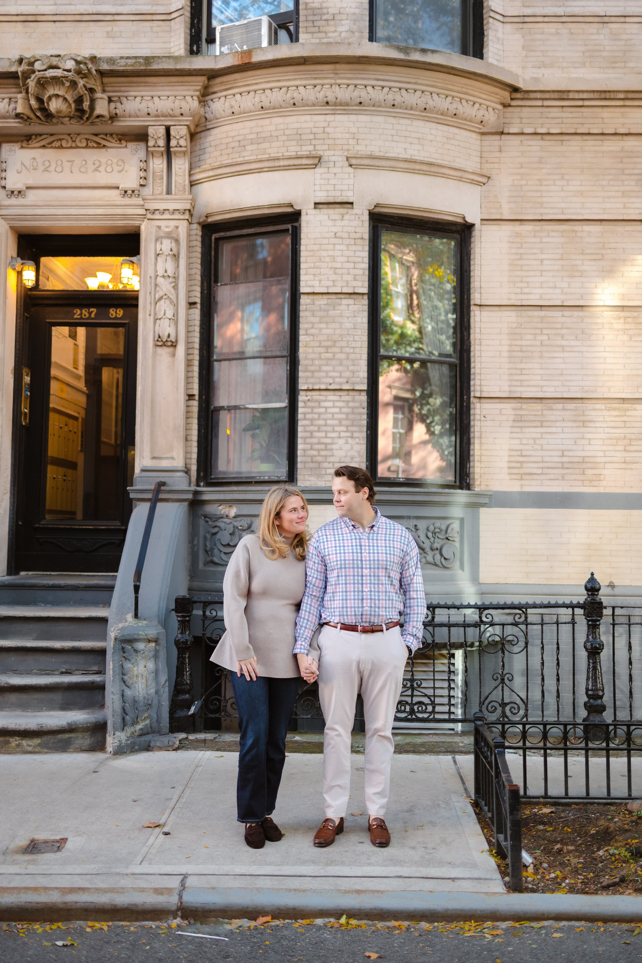 A couple holding hands stands on a sidewalk in front of a beige brick building with large windows and a black door.