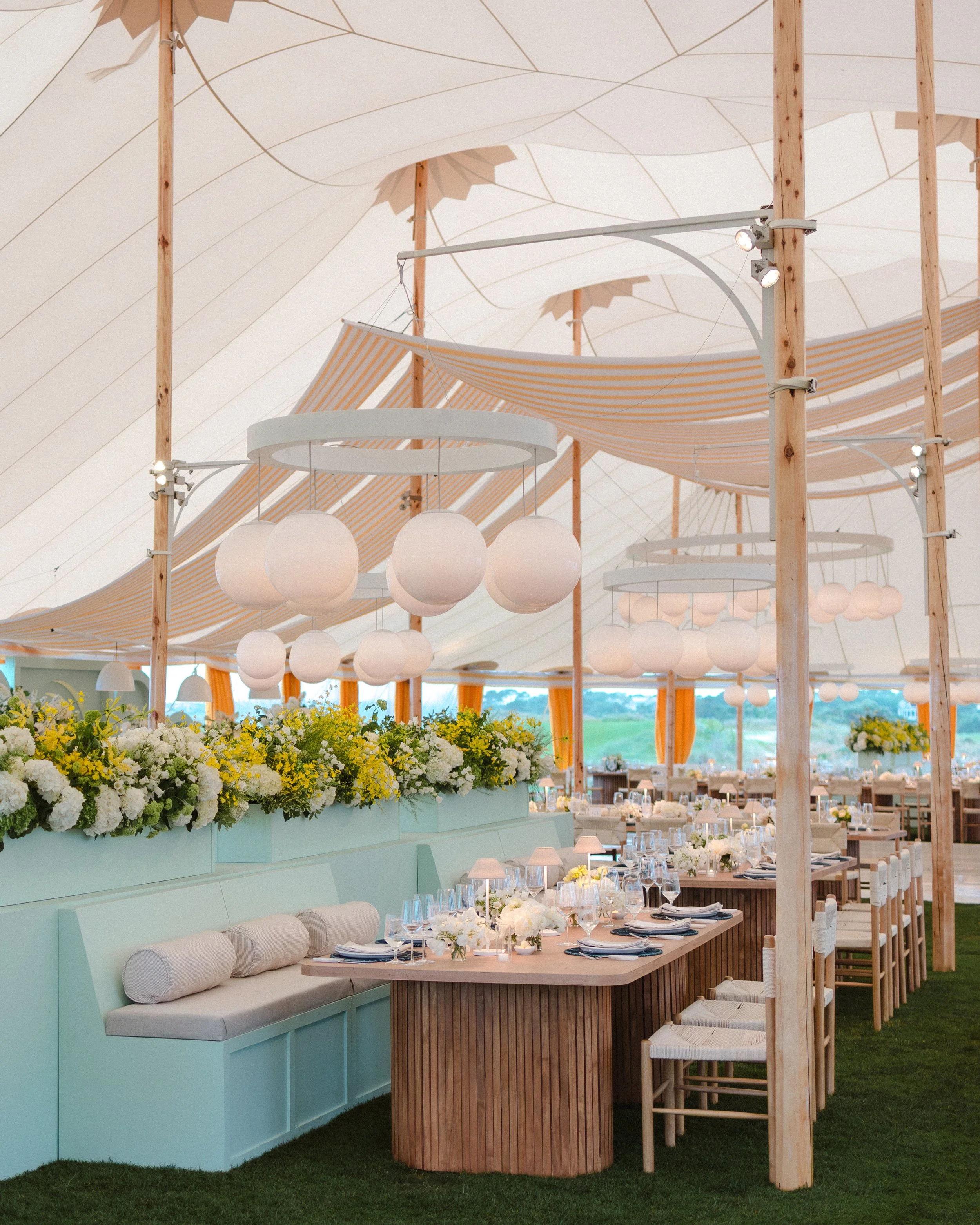 Elegant outdoor wedding reception under a large white tent with modern decor, round white paper lanterns, and floral arrangements with white and yellow flowers. Designed by Reagan Events