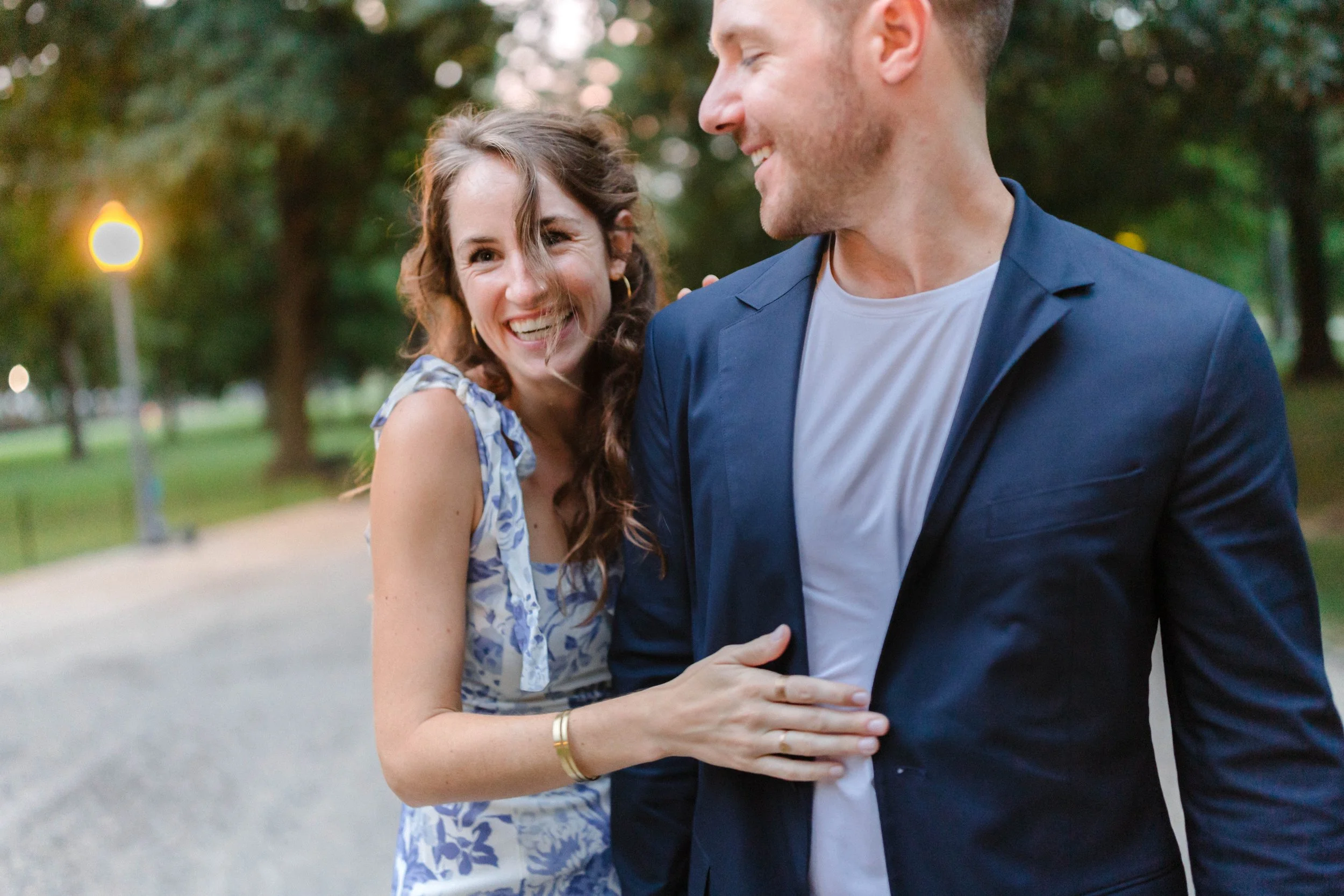 A woman and a man smiling and laughing outdoors on a park pathway, with trees and streetlights in the background. The woman is wearing a blue and white floral dress, and the man is wearing a navy blazer over a white t-shirt.