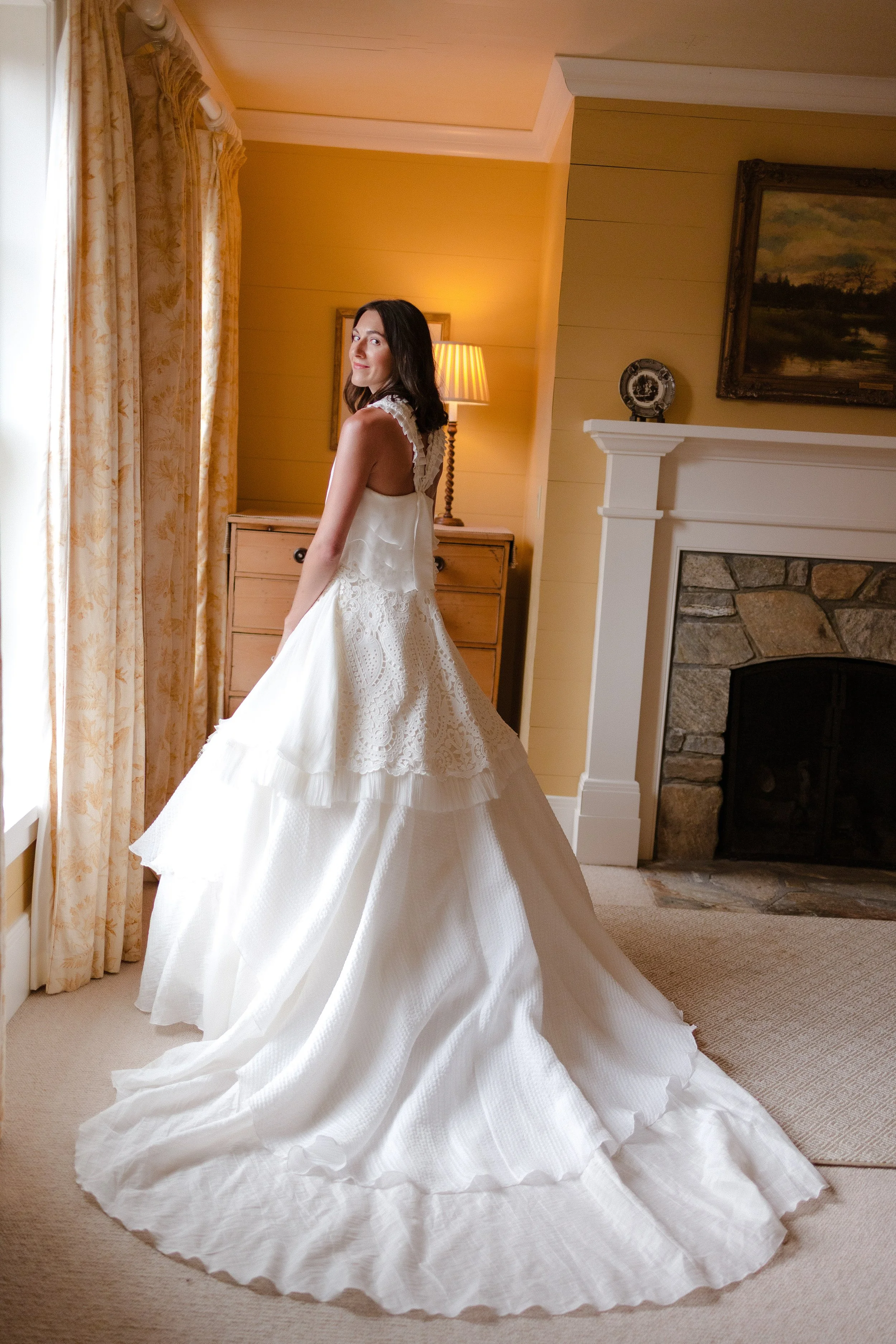 A woman in a white wedding gown standing in a warmly lit room with a fireplace and a window with floral curtains.