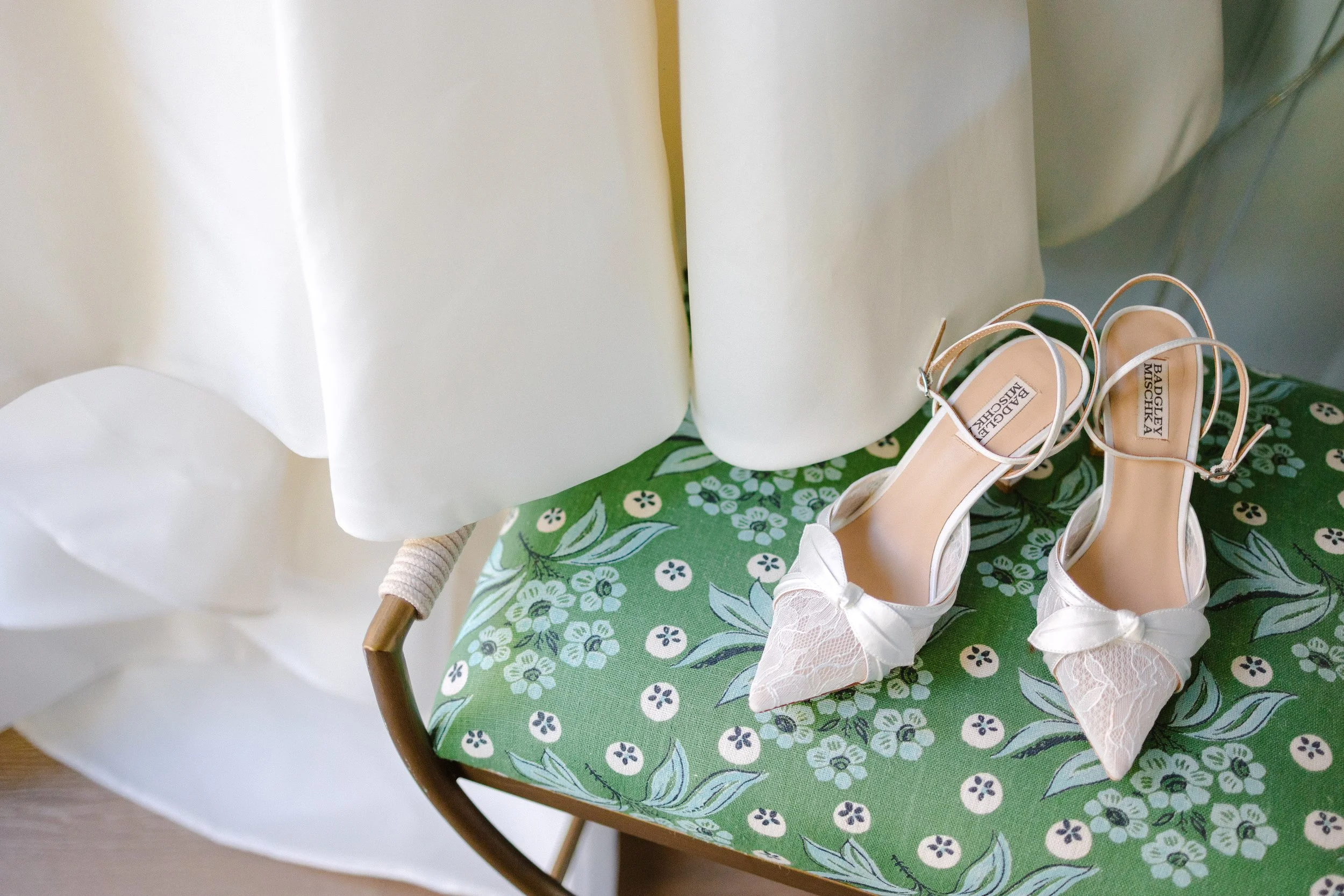 A pair of white high-heeled wedding shoes with lace details and bows, placed on a floral green fabric surface, beside a white dress hanging nearby.