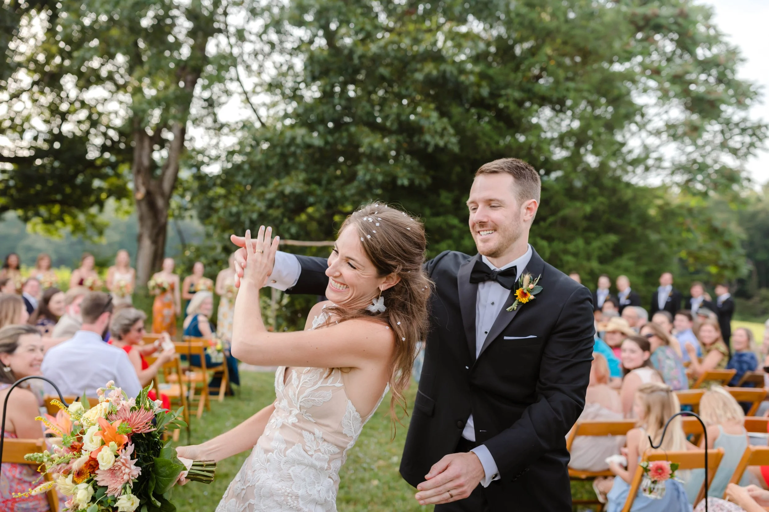 A joyful bride and groom dancing at their outdoor wedding ceremony, with guests seated on wooden chairs and a lush green background.