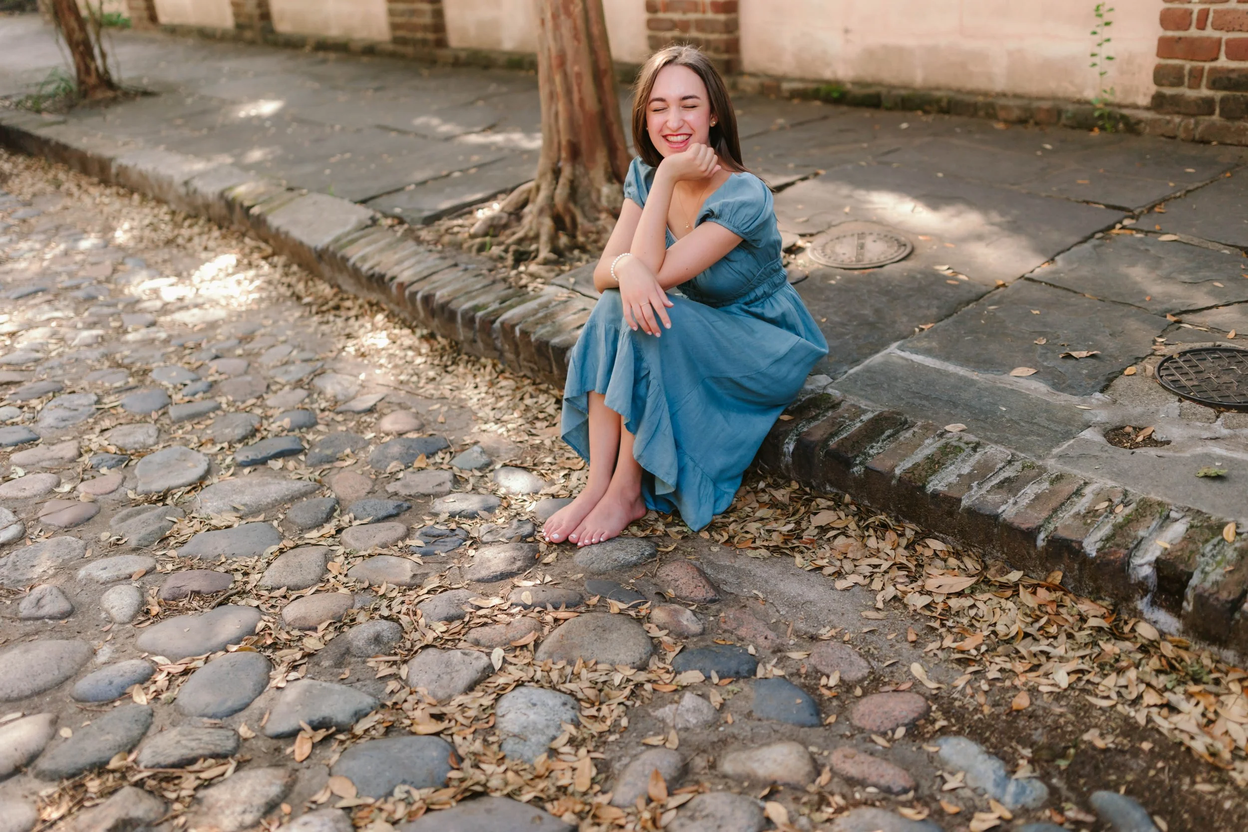 A woman sitting on a sidewalk with cobblestones and fallen leaves, smiling with eyes closed, wearing a blue dress, in a setting with brick walls and a tree.