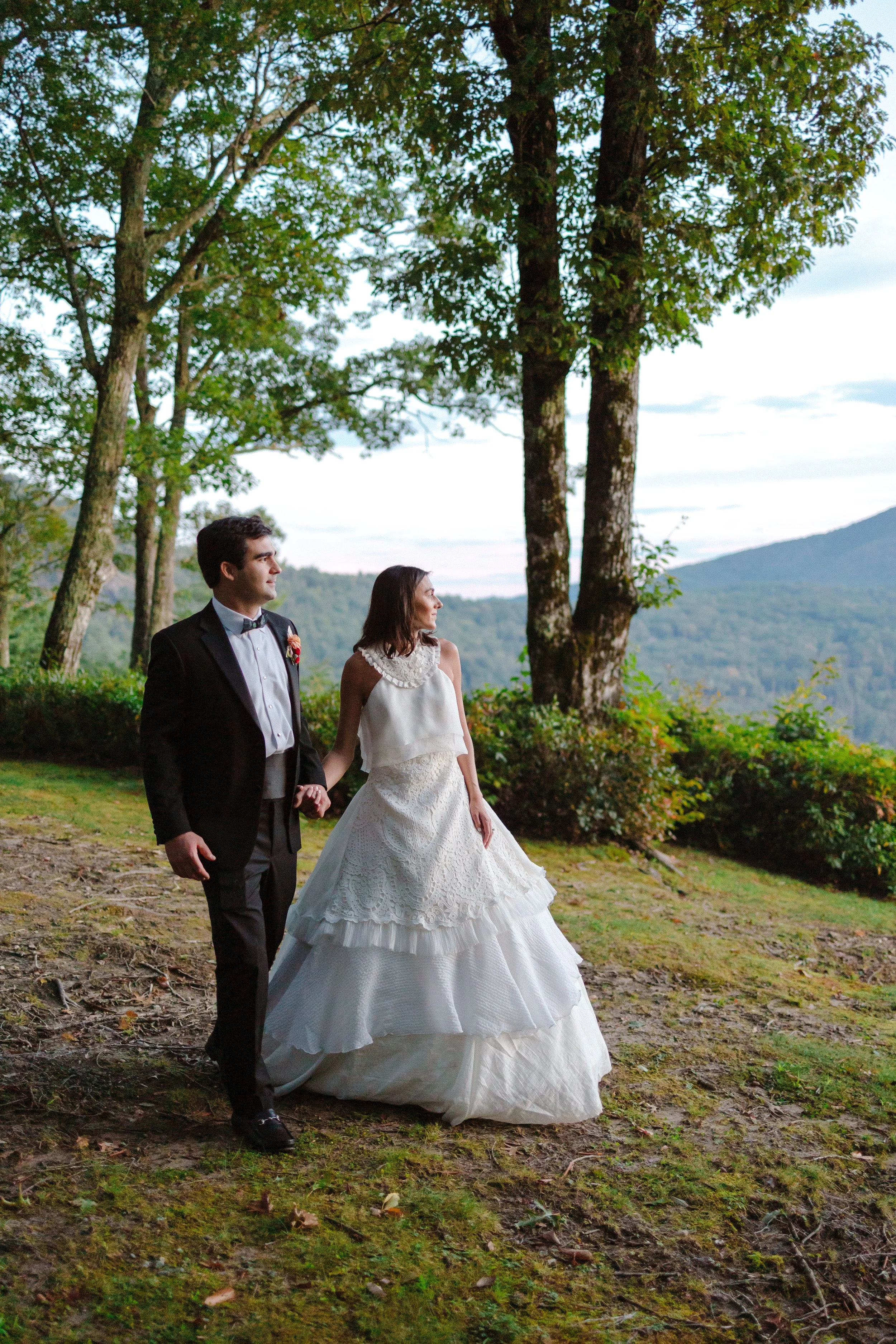 A bride and groom walking outdoors in a wooded area on their wedding day, holding hands, with mountainous landscape in the background.